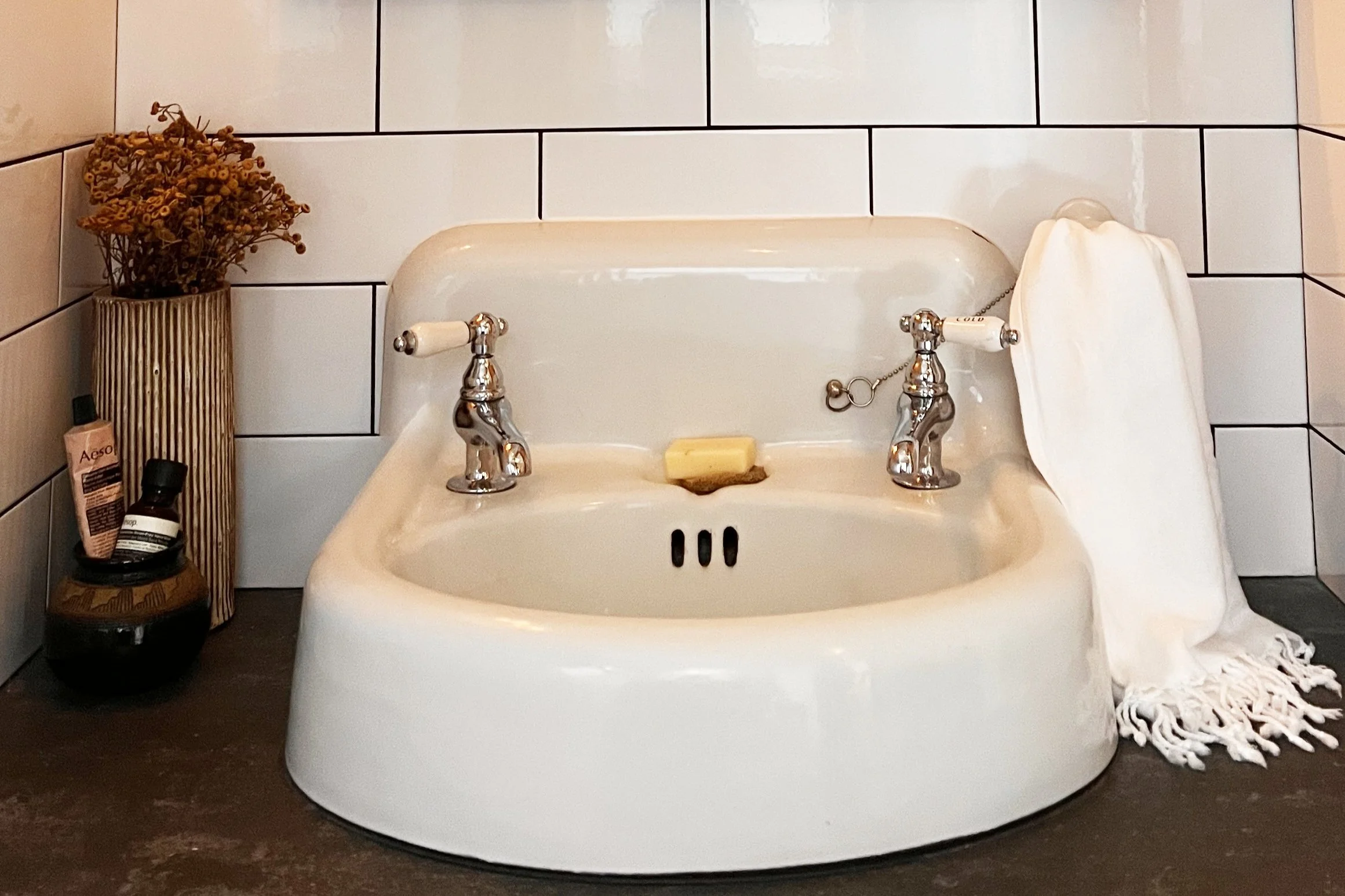 A vintage-style wall-mounted sink with a rounded basin and separate chrome taps for hot and cold water sits against a backdrop of white subway tiles with dark grout. To the left of the sink, a black dish holds personal care products and a ribbed vase