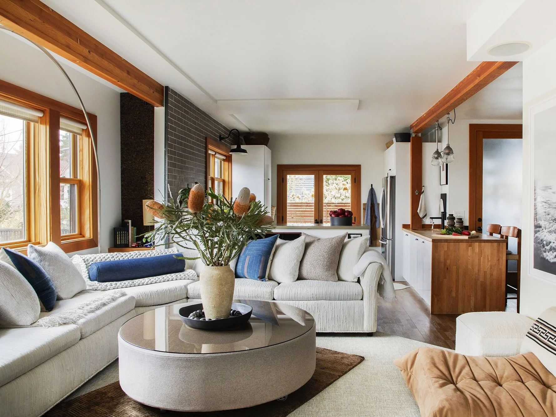 A modern, open-concept living room and kitchen with natural wood accents and large windows letting in ample daylight. The space features a white sectional sofa with blue and gray throw pillows, a round beige ottoman-style coffee table with a vase of 
