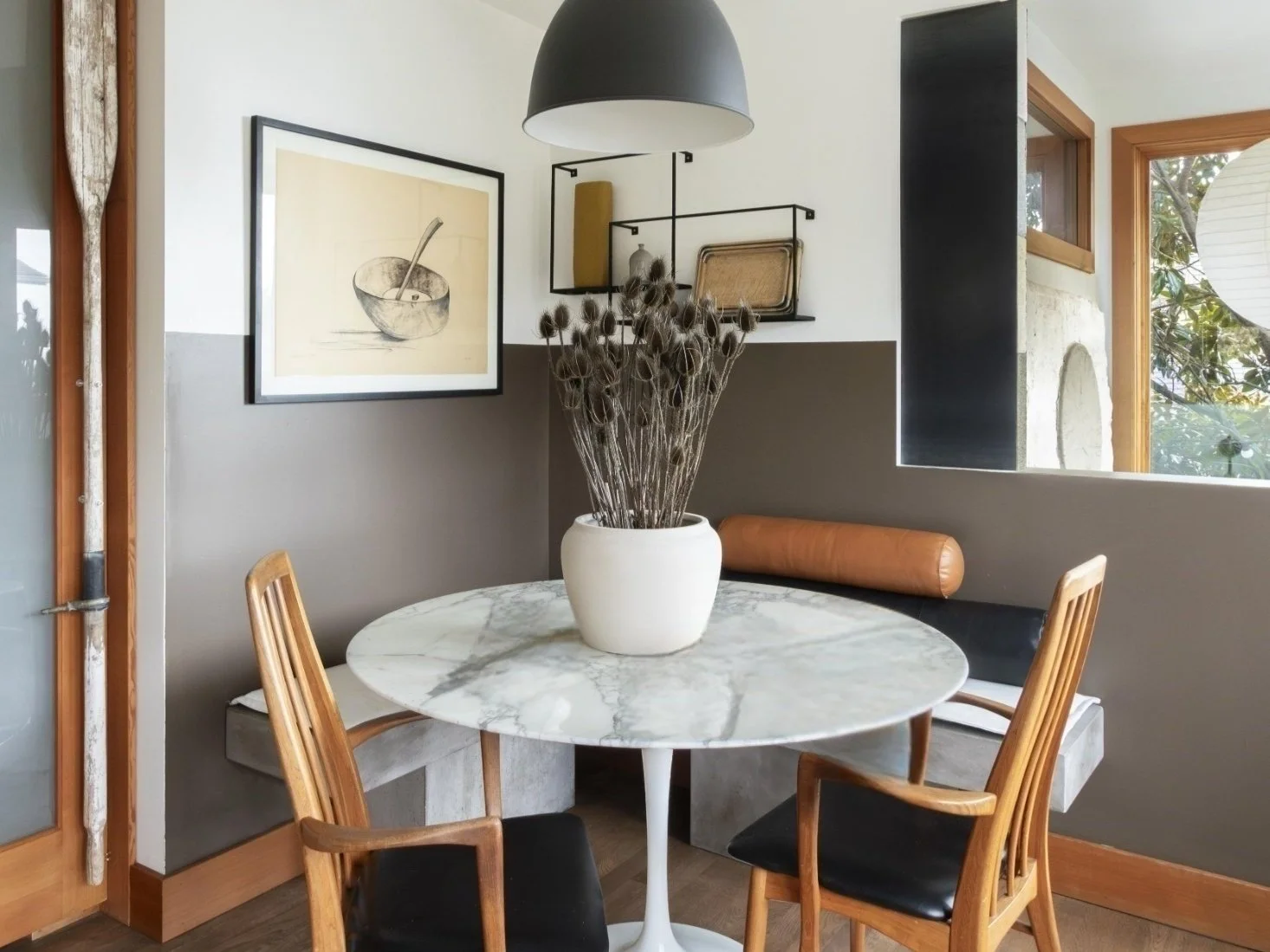 A cozy and modern dining nook featuring a round white marble table surrounded by a mix of built-in bench seating and wooden chairs with black cushions. A large ceramic vase with dried floral stems serves as the centerpiece. The walls are painted in t