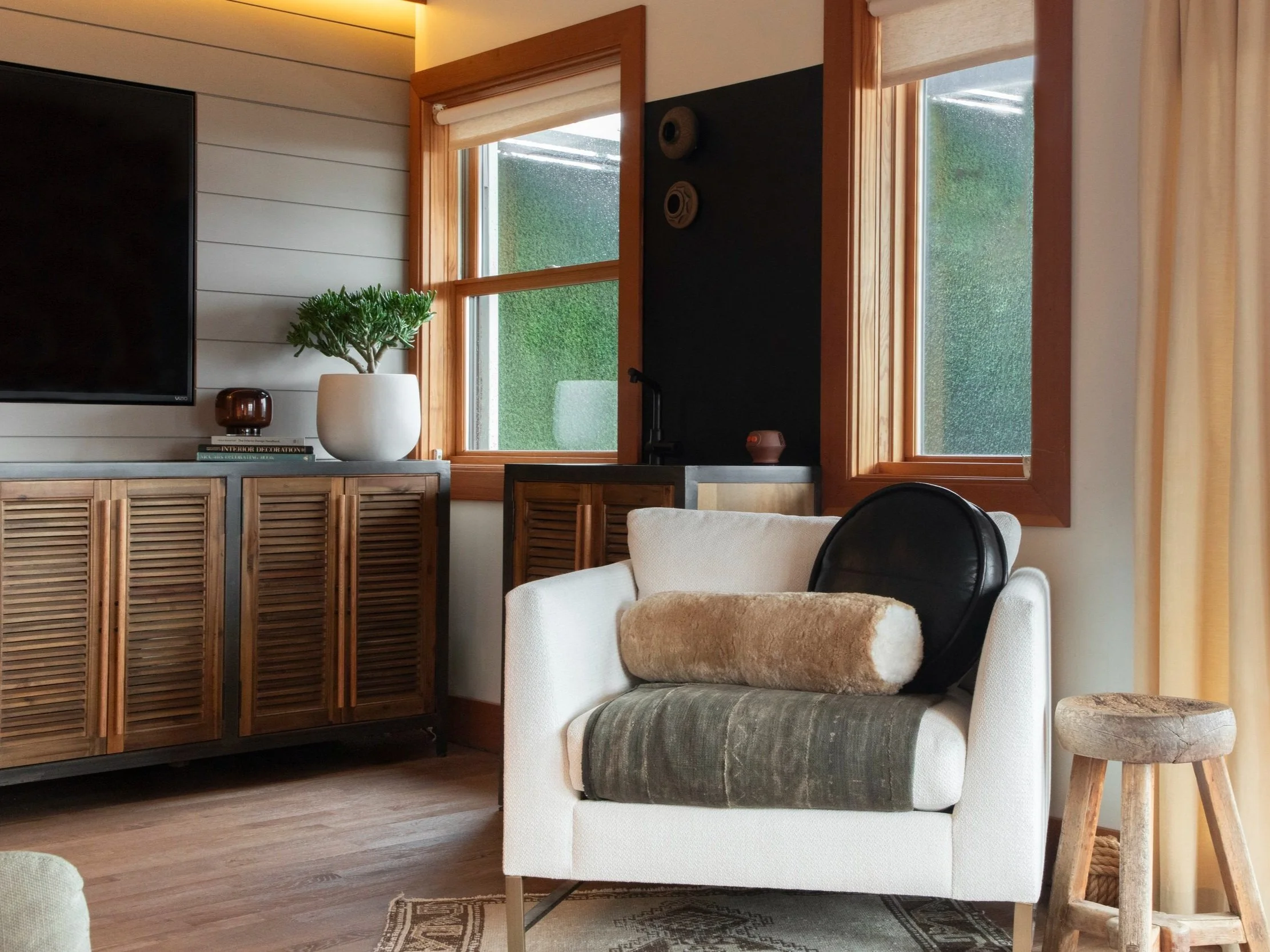 A cozy, well-lit living room corner featuring a modern white armchair layered with textured pillows and a soft green throw. The chair is positioned beside a small rustic wooden stool and set against a backdrop of large windows with natural wood frame