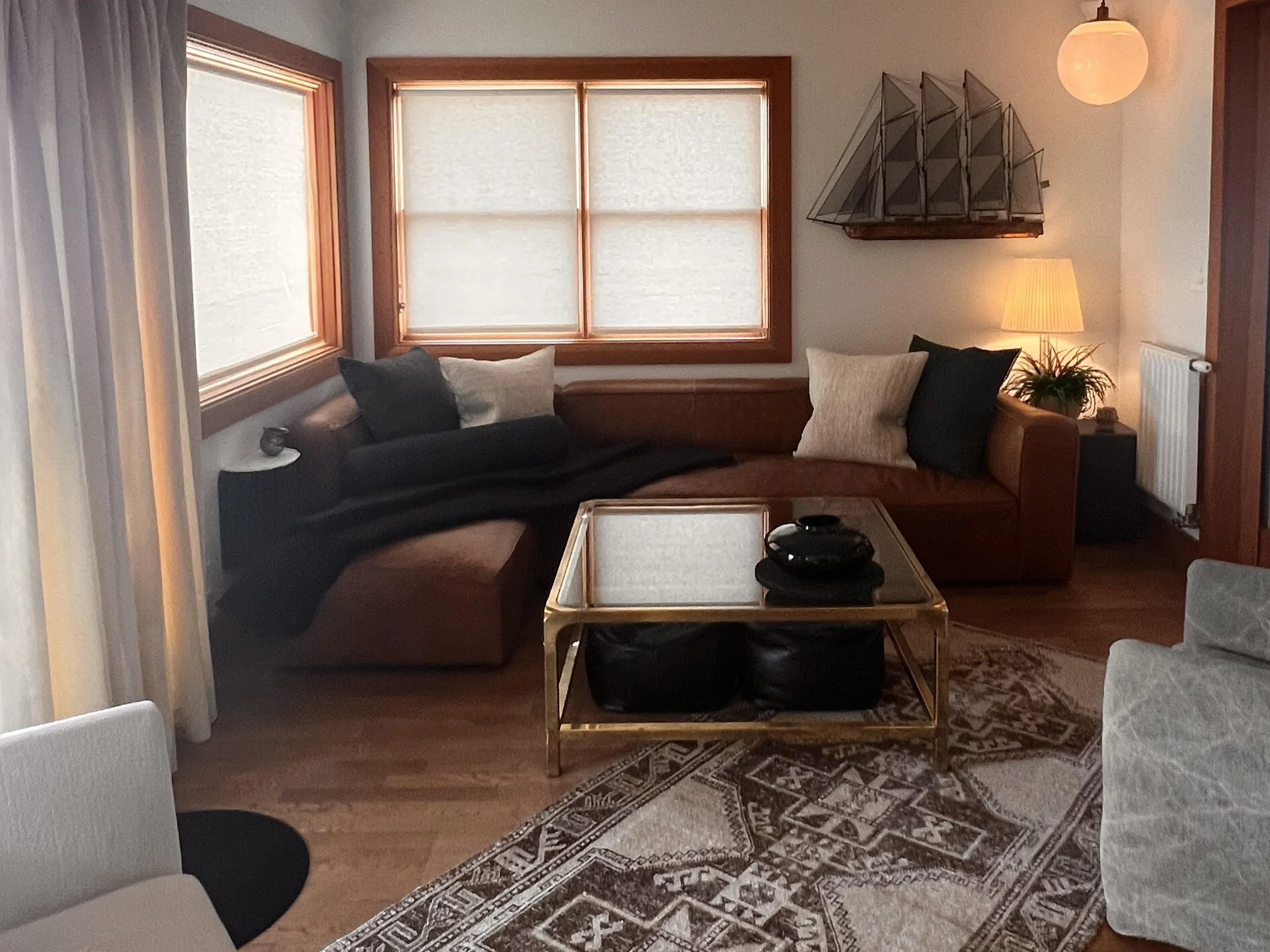 A cozy and stylish living room corner with a large L-shaped brown leather sofa accented by a mix of neutral throw pillows and dark blankets. The space is warmly lit by natural light through two large windows with woven blinds, and a soft-glow table l