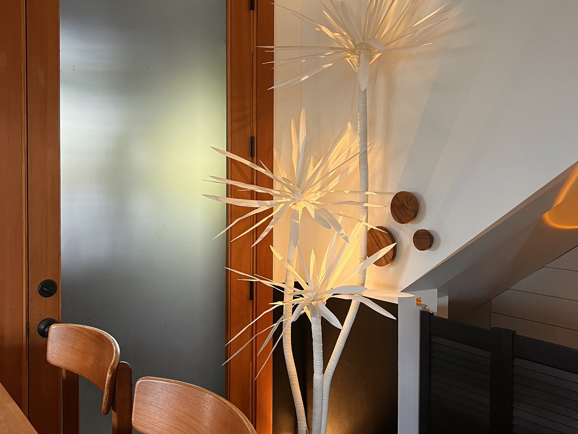 A warmly lit interior corner showcasing a sculptural, white floor lamp shaped like a spiky-leafed plant, casting dramatic shadows on the adjacent wall. The lamp stands next to a frosted glass door framed in natural wood, adding to the soft, ambient g