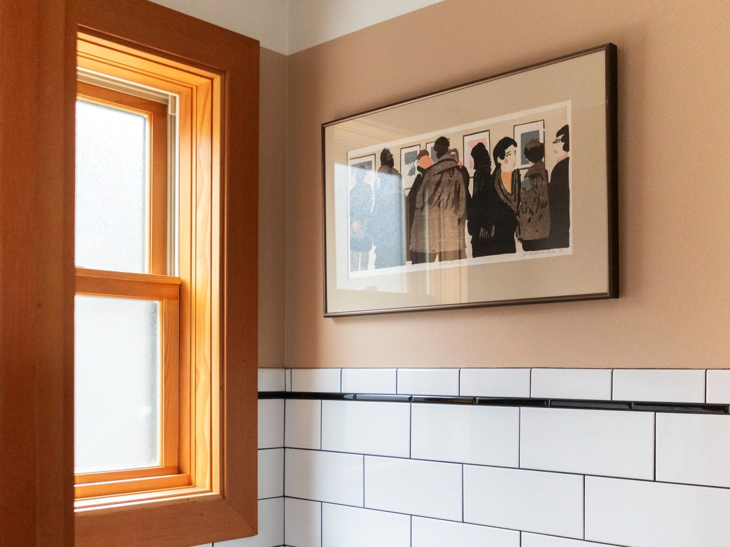 A small, stylish room with white subway tiles and a beige upper wall features a wooden-framed window that lets in natural light. Above the tile line, a framed piece of artwork depicting a group of people viewing images in a gallery adds visual intere