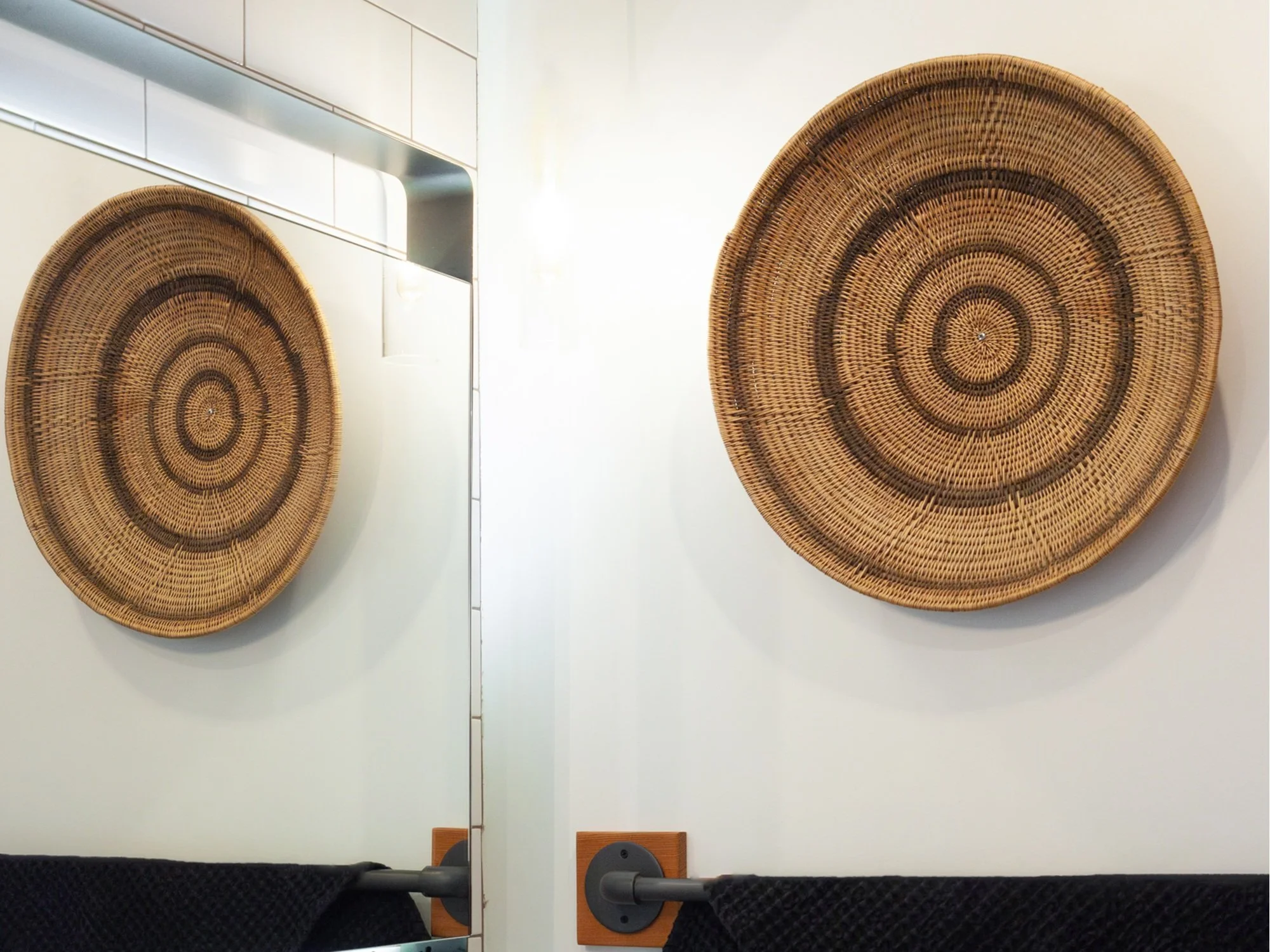 A close-up of a bathroom wall featuring a large woven basket hung as wall decor. Its concentric circular pattern is mirrored in the adjacent wall mirror, creating visual symmetry. A dark textured towel hangs on a matte black towel bar beneath the bas