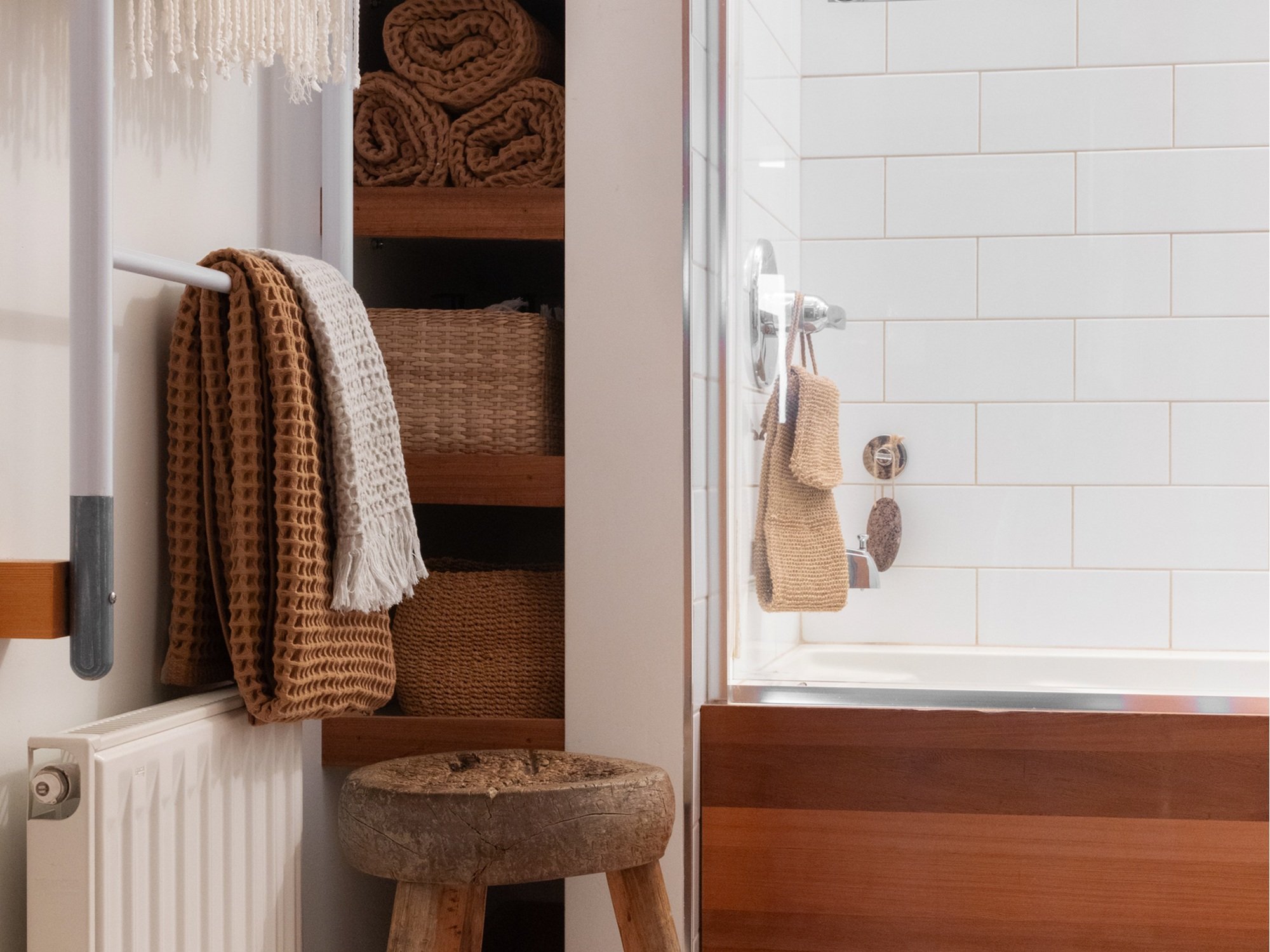 A cozy, well-organized bathroom featuring earthy tones and natural textures. On the left, open wooden shelves are neatly stacked with rolled and folded waffle-knit towels and woven baskets. A wooden stool with a rustic finish sits in front, and a rad