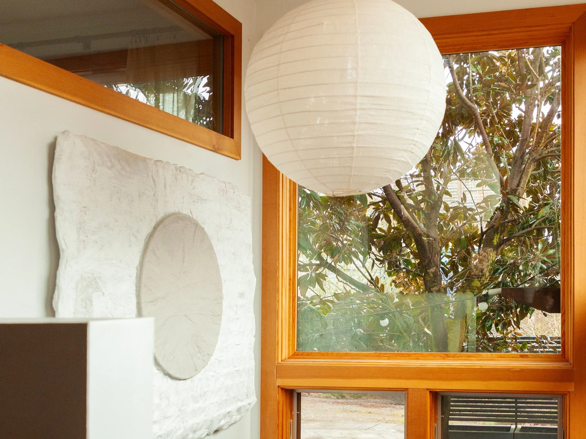 A serene corner filled with natural light from large windows framed in warm wood, offering a view of a leafy tree outside. A white paper lantern-style pendant light hangs from the ceiling, and a textured white wall sculpture with a circular design ad