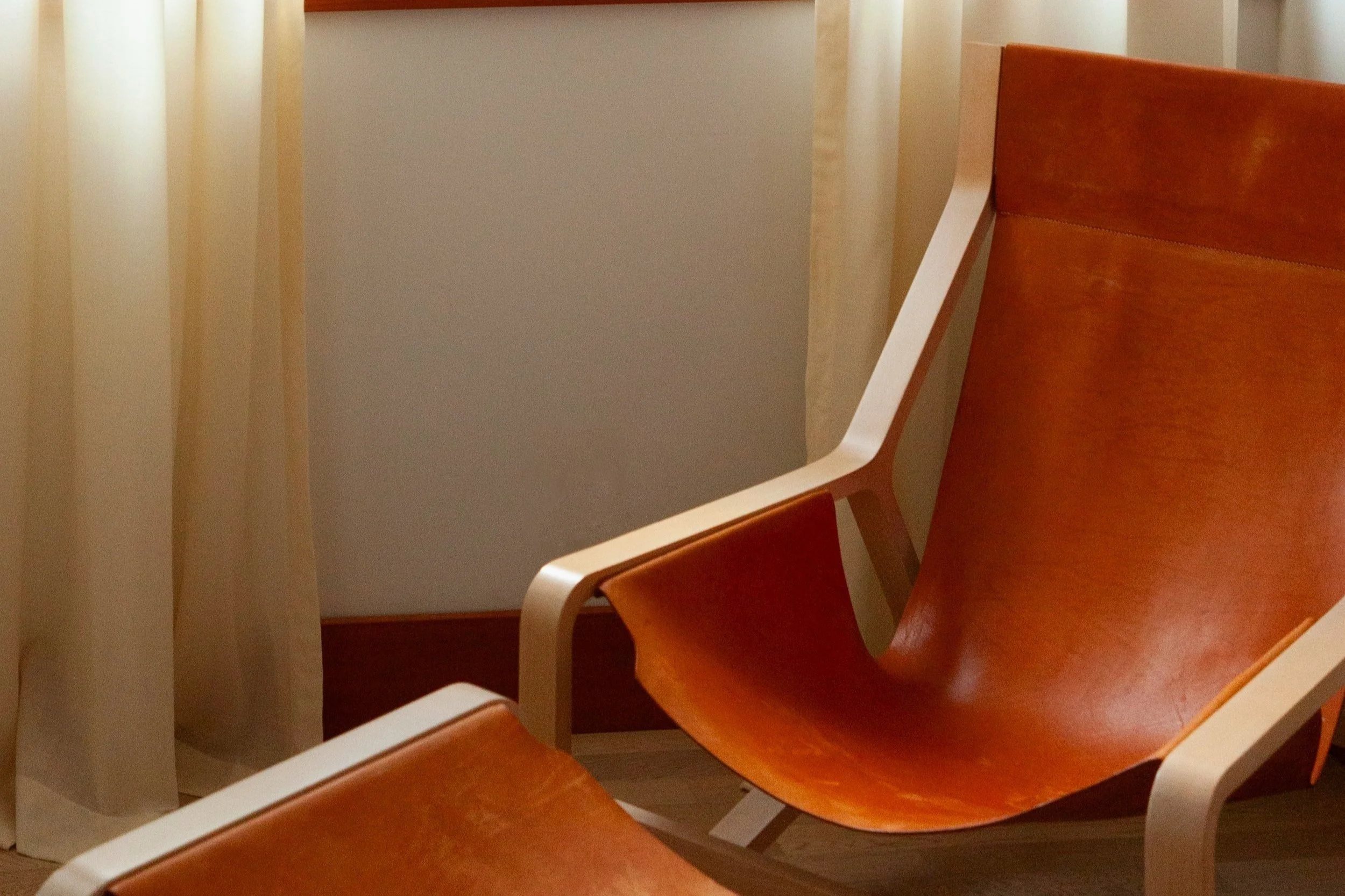 A close-up view of two minimalist lounge chairs with light wooden frames and rich, cognac-colored leather slings, set against a backdrop of sheer cream curtains and a neutral wall. The soft natural light filtering through the curtains enhances the wa