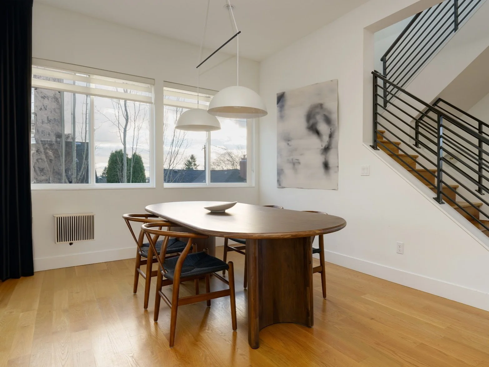 A modern dining area with a minimalist and sophisticated design. The space features a dark wood oval dining table surrounded by matching wooden chairs with black seat cushions. Above the table hang two oversized white dome pendant lights, adding scul