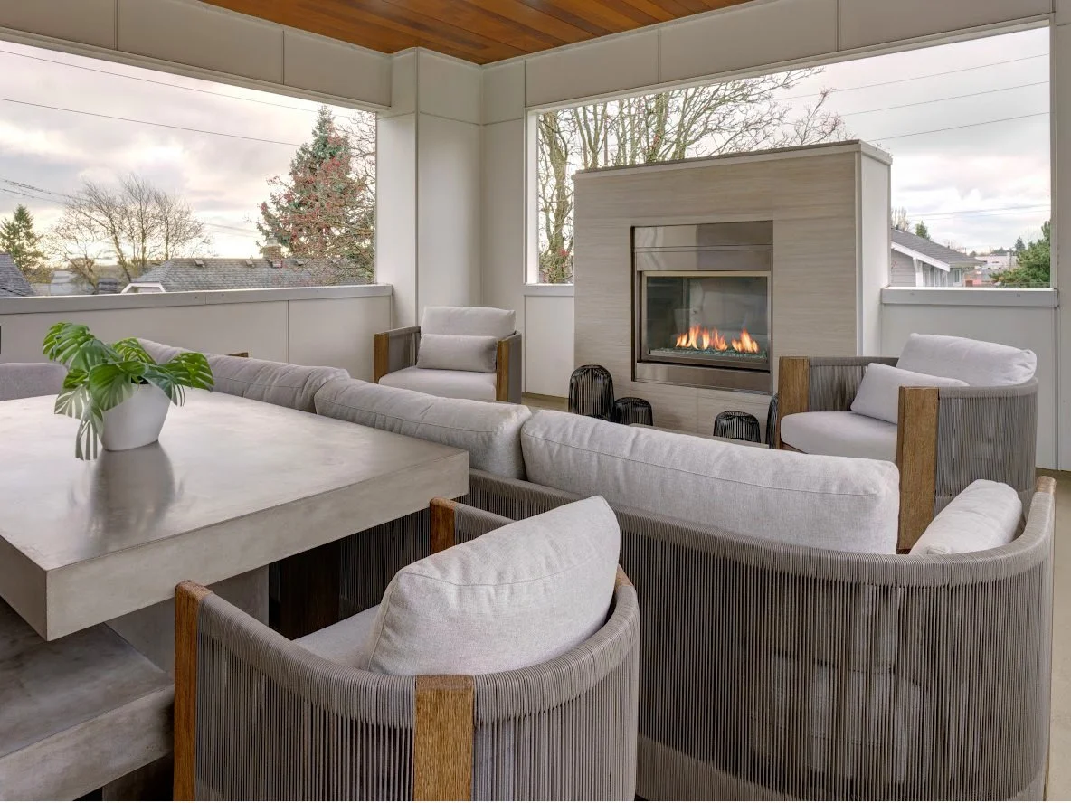 A stylish and serene covered outdoor living space designed for comfort and relaxation. The area features a modern gas fireplace set into a sleek, stone-look surround, providing warmth and ambiance. Plush, light-gray cushioned seating—including armcha