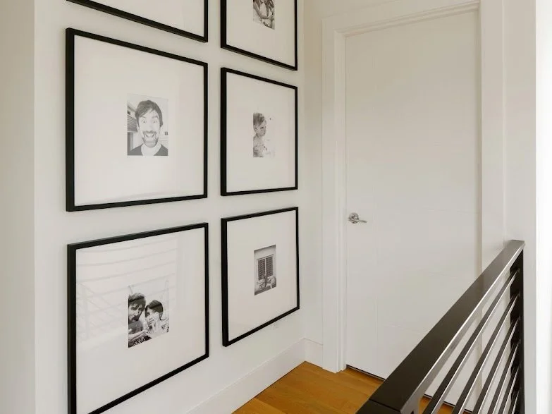 A modern hallway with a clean and minimalist aesthetic, featuring a gallery wall of six black-and-white photographs arranged in a grid. Each image is matted and framed in simple black frames, creating a cohesive and elegant display. The space is defi