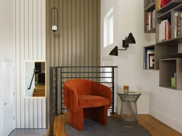 A cozy and modern reading nook tucked into a stair landing, featuring a bold burnt-orange velvet armchair placed on a soft gray rug. Beside the chair is a stylish round side table with a wire metal base and glass top, holding a small candle. A black 
