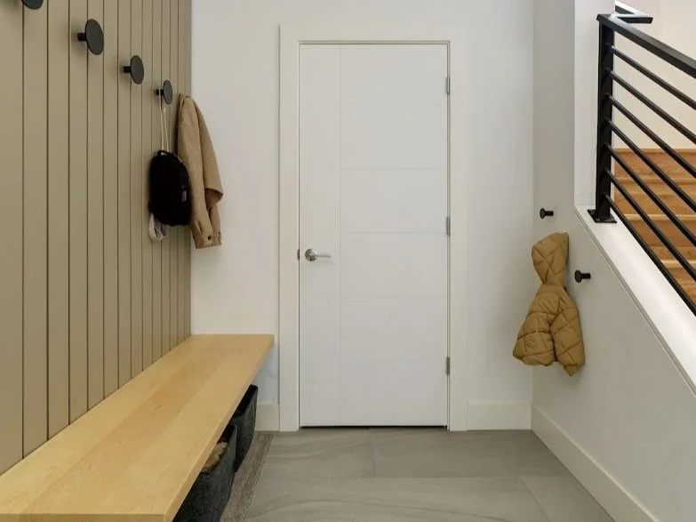 A sleek and functional mudroom or entryway with a minimalist design. On the left, vertical olive-colored slats with black circular wall hooks offer hanging space for coats and bags, one of each currently displayed. Below is a light wood bench with cu