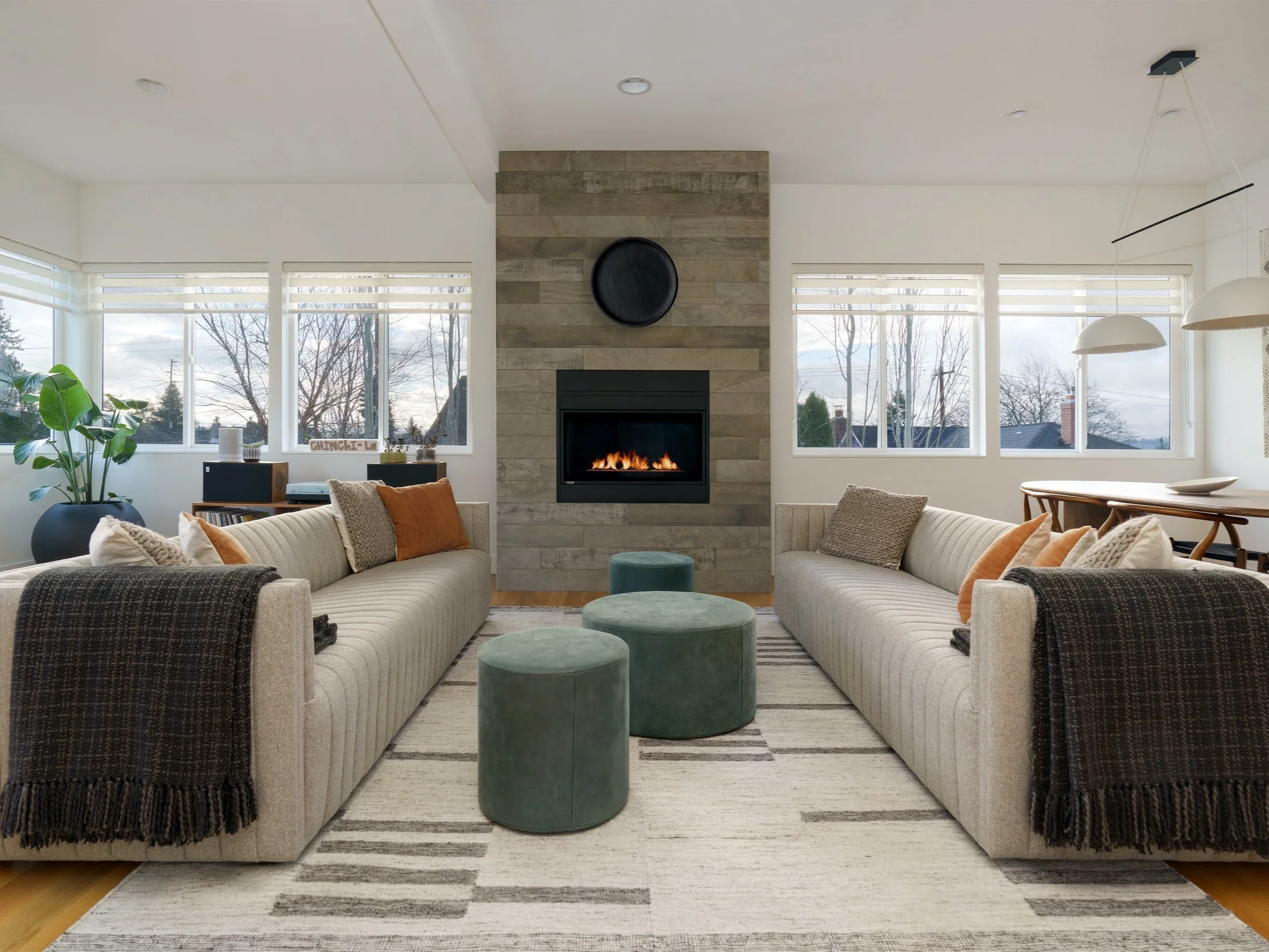 A modern and cozy living room with a symmetrical layout centered around a sleek, tiled fireplace with a built-in gas insert. Two identical light beige sofas with vertical channel tufting face each other, adorned with a mix of neutral and rust-colored