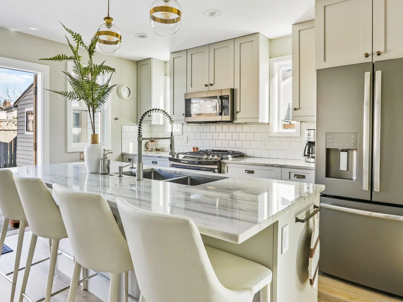 A bright, contemporary kitchen with a fresh, inviting aesthetic. The space features a striking marble-topped island with a waterfall edge and an undermount sink, accompanied by four sleek white bar stools. Above the island hang two modern pendant lig