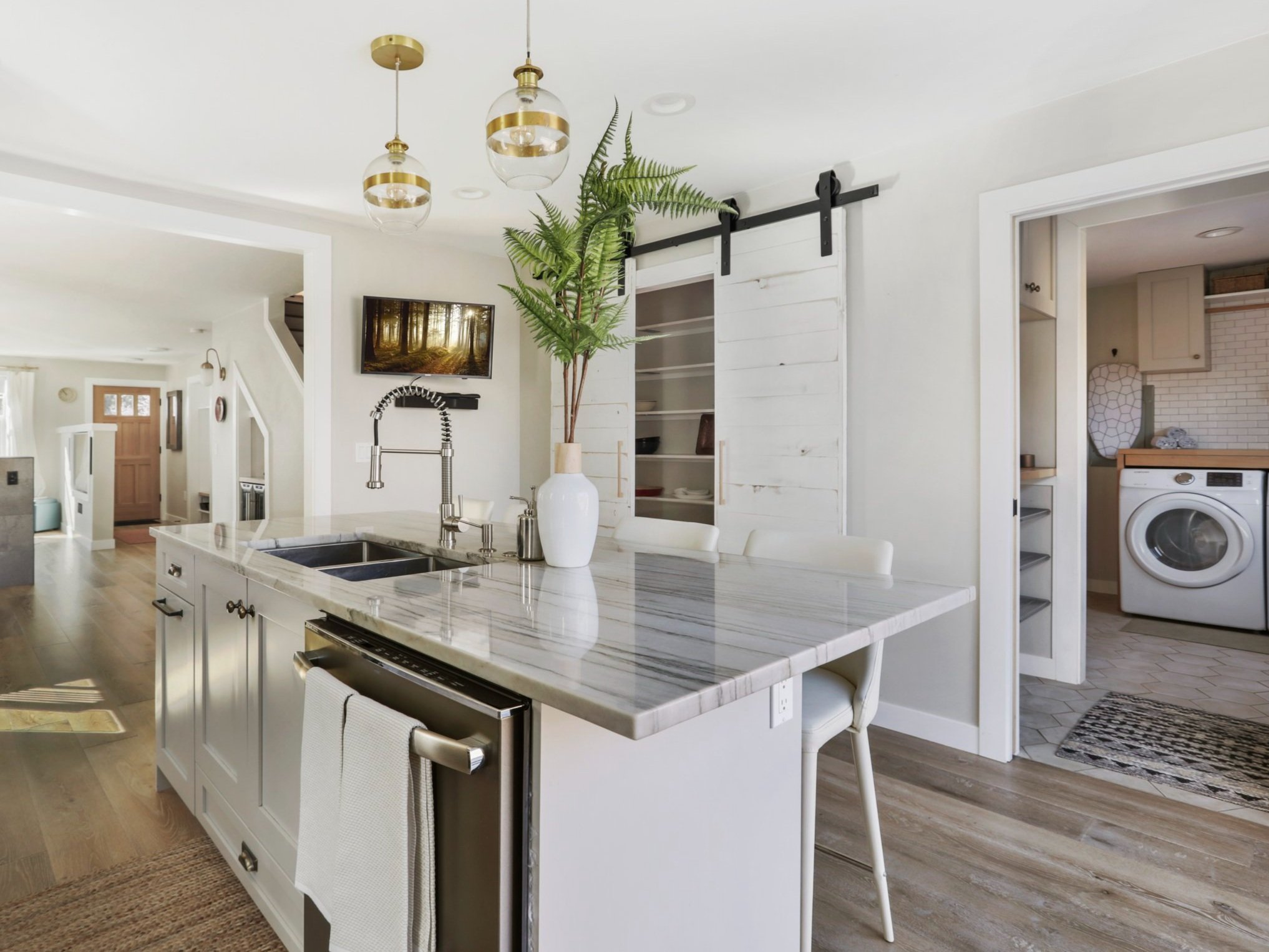 A bright, modern kitchen featuring a central island with a marble waterfall countertop, integrated sink, and built-in dishwasher. The island is accompanied by white upholstered bar stools and decorated with a tall white vase of greenery. Above the is