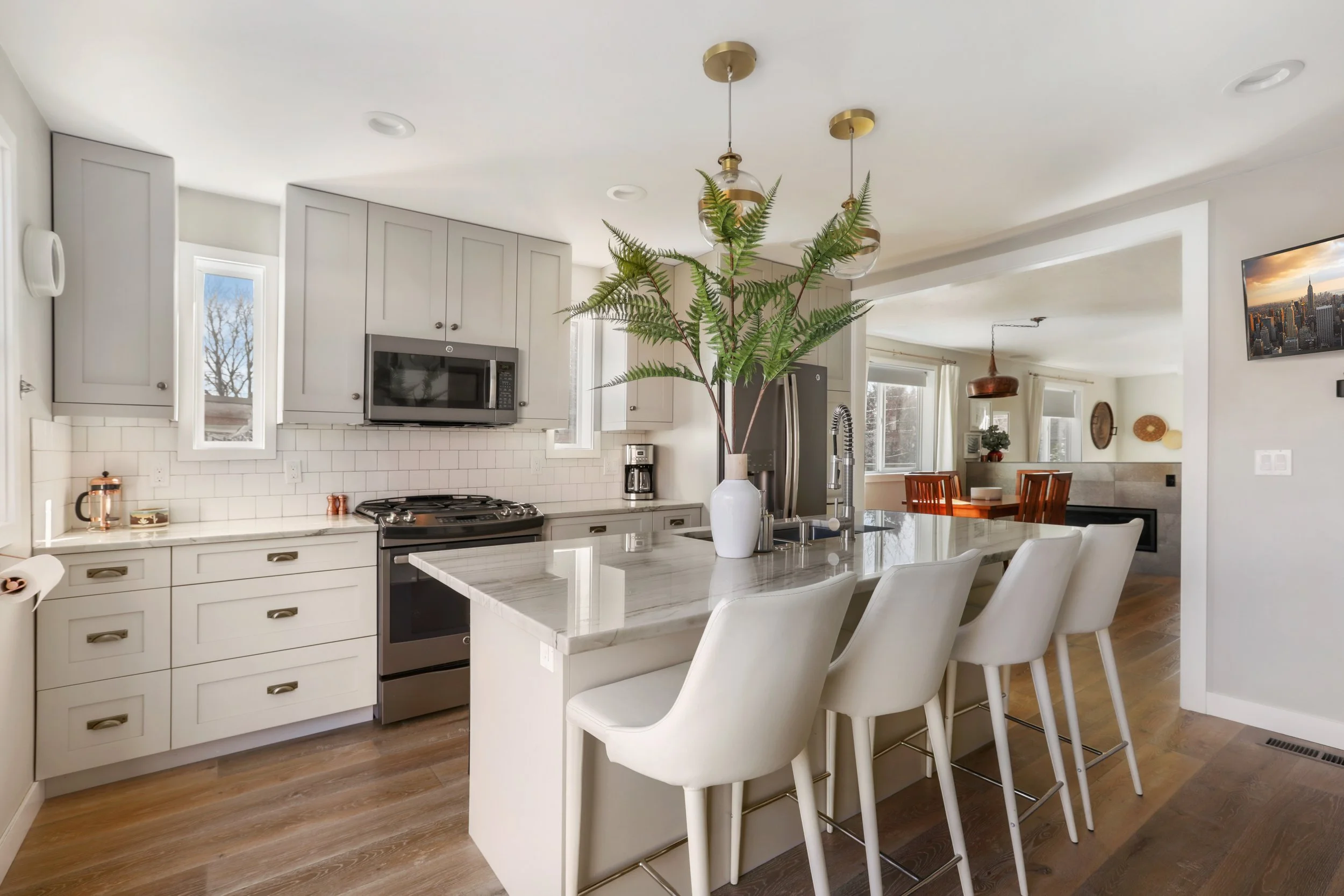 A stylish and open-concept kitchen with a central island featuring a waterfall-edge marble countertop and four white upholstered bar stools. The kitchen cabinetry is a soft gray, paired with a white subway tile backsplash and stainless steel applianc