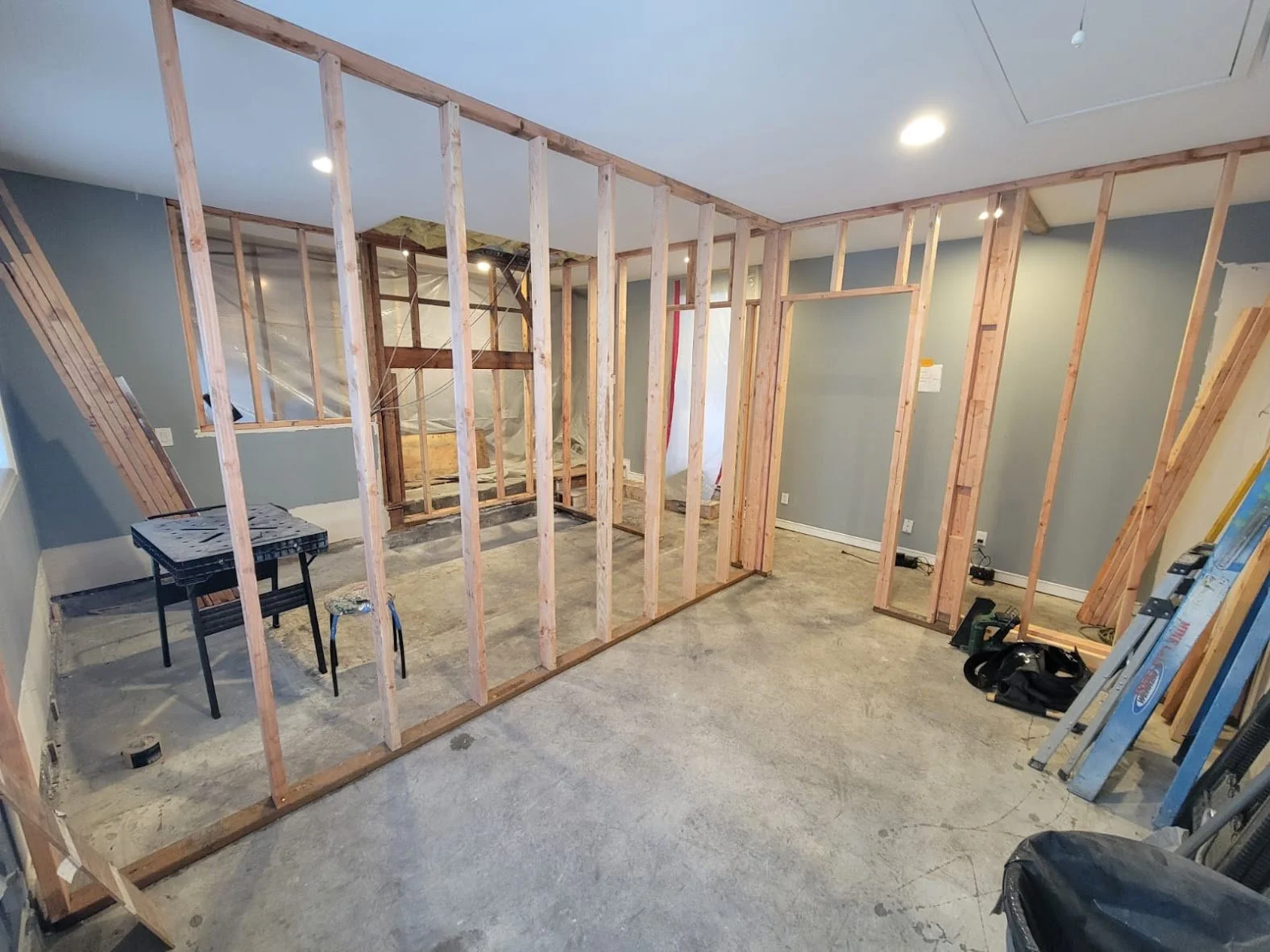 This image shows a residential interior under construction, with wood framing in place for new walls, indicating a renovation or remodeling project in progress. The floor is bare concrete, and the studs have been installed to define separate rooms, i