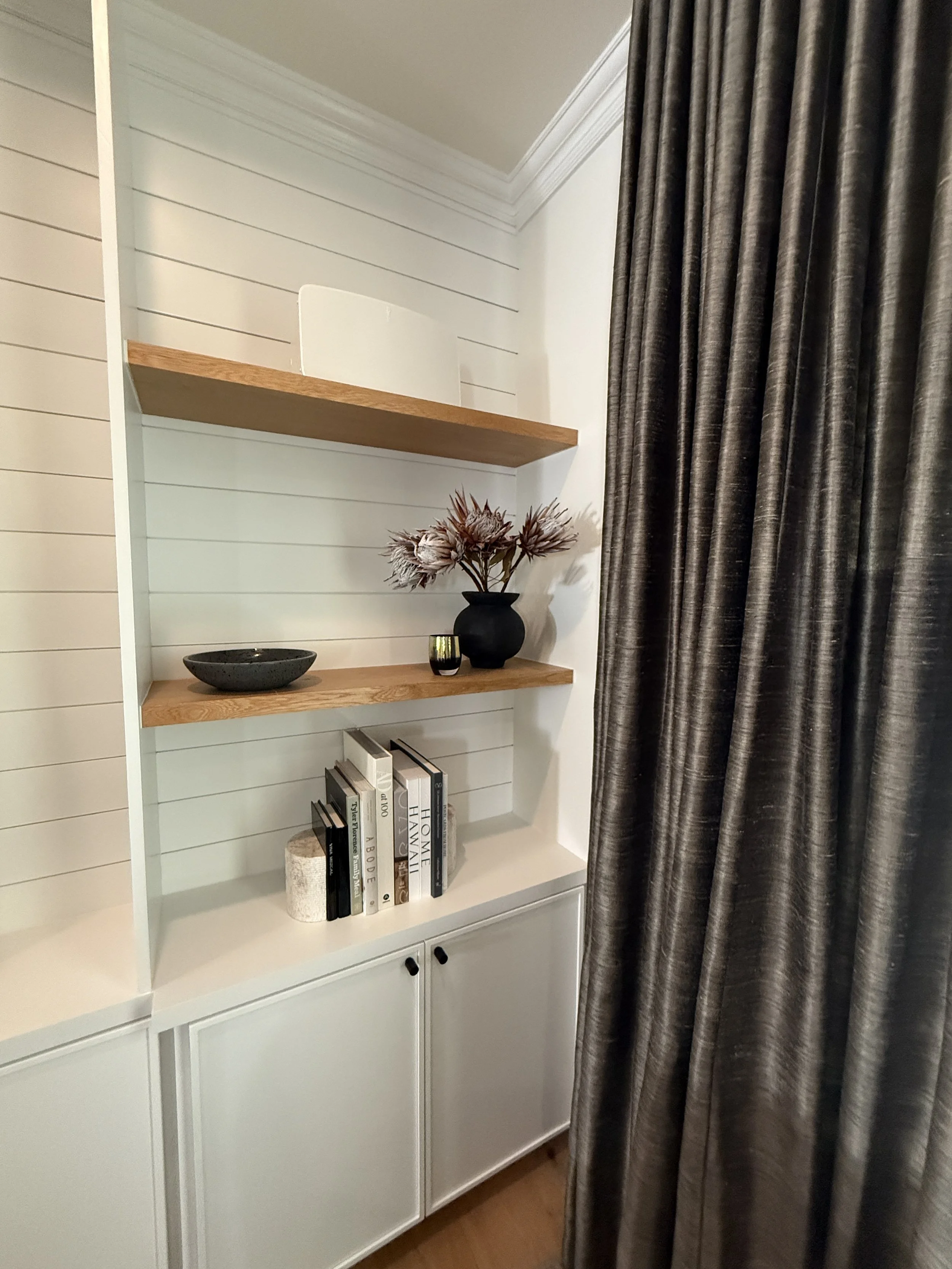 A close-up of a stylish built-in shelving nook, featuring white shiplap walls and two natural wood floating shelves. The top shelf displays a minimal white decor piece, while the lower shelf holds a black vase with dark, wispy foliage and a few stack