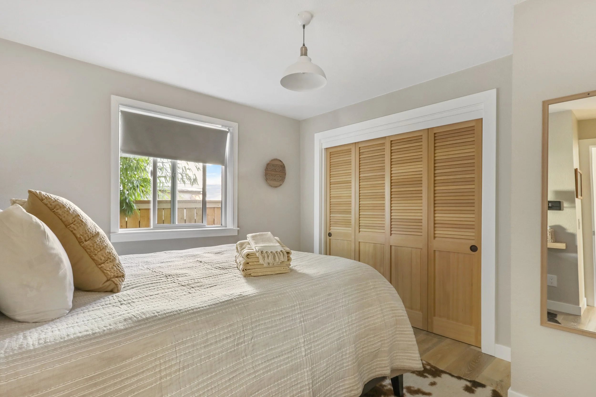 A bright and serene bedroom decorated in soft, neutral tones. The bed is dressed in beige and cream bedding with neatly arranged pillows and folded towels at the foot. A large window with a gray roller shade lets in natural light, while wooden louver