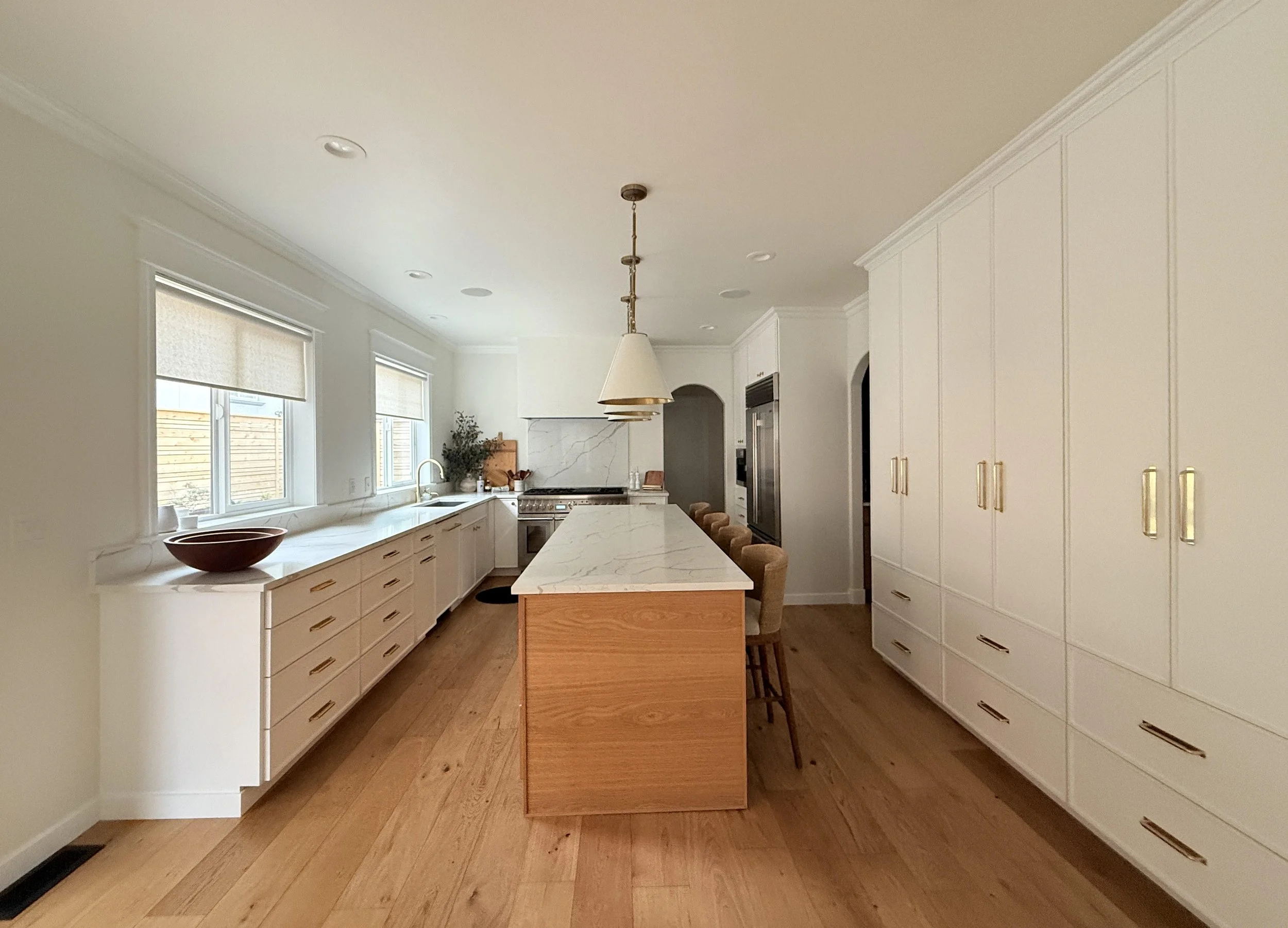 A spacious, light-filled modern kitchen with a clean and streamlined aesthetic. The design features a large central island with a wood base and a white marble countertop, surrounded by woven barstools. On one side, there’s an extended run of white ca