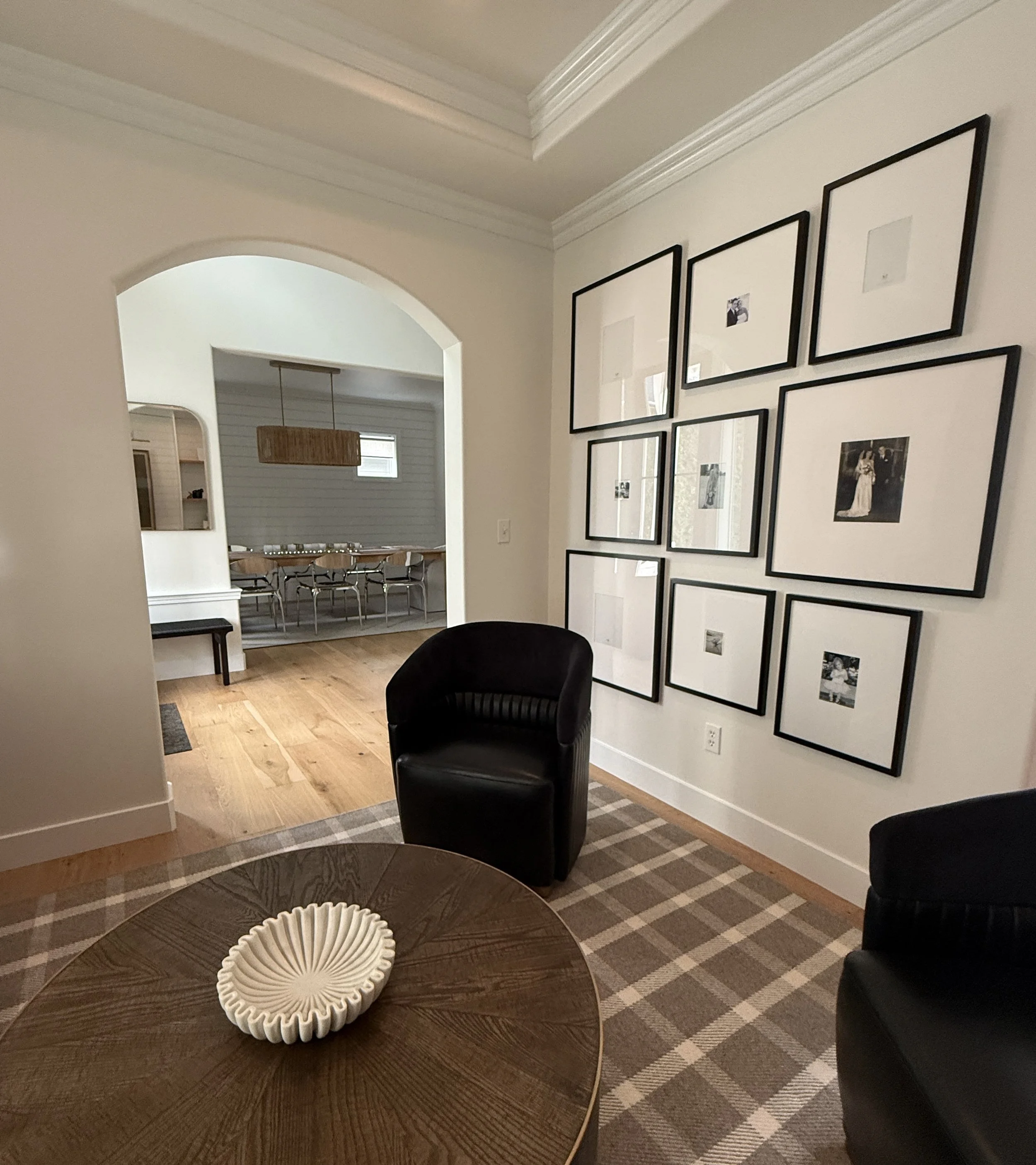 A stylish transitional space featuring a small sitting area with two black barrel chairs and a round wooden coffee table topped with a sculptural white bowl. The floor is covered in a brown plaid-patterned rug, adding a cozy and classic touch. A gall