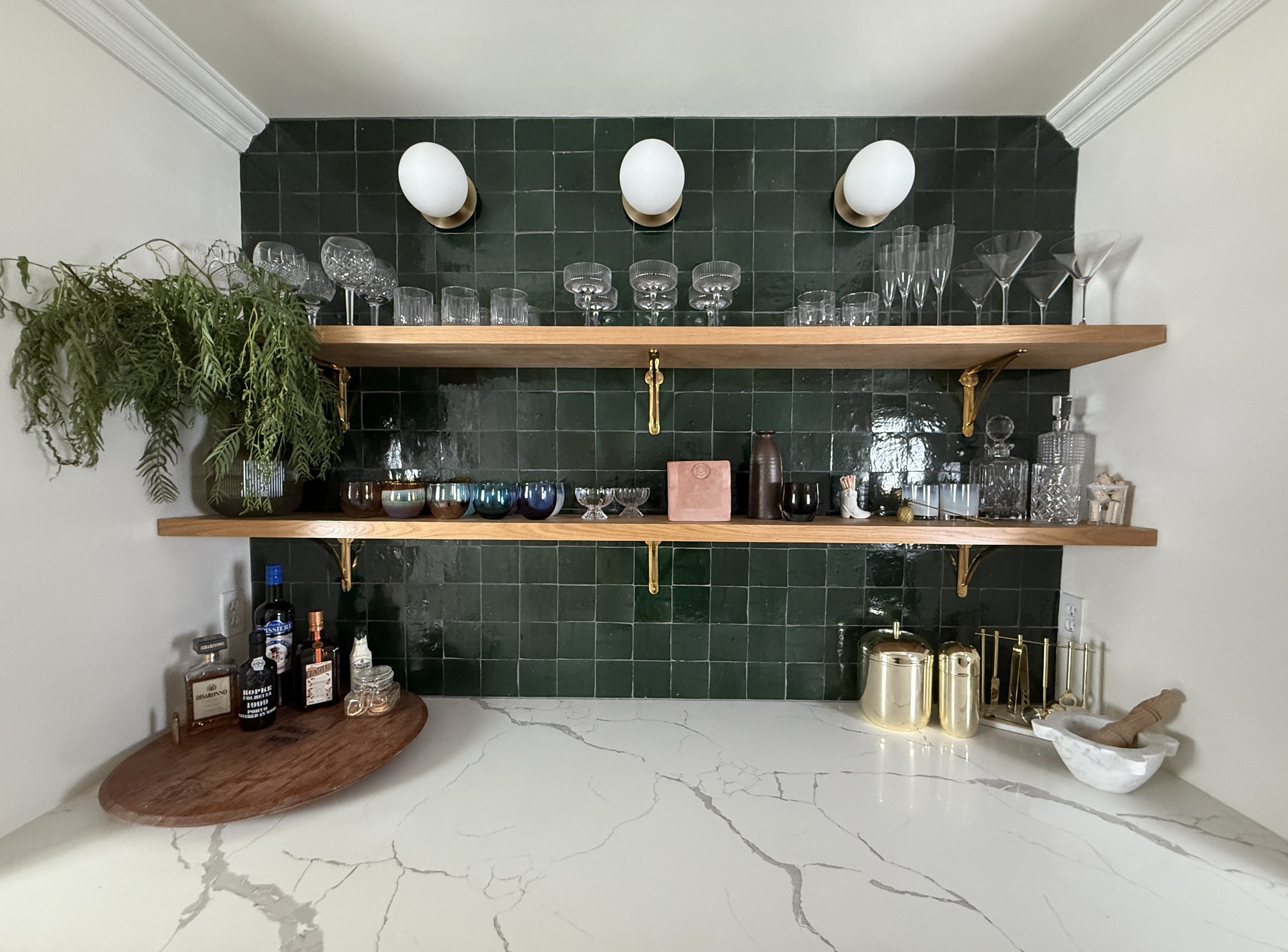 A chic and well-organized home bar setup featuring a dark green tiled backsplash that adds dramatic contrast to the white marble countertop. Two wooden floating shelves with gold brackets hold an assortment of elegant glassware, including wine, cockt