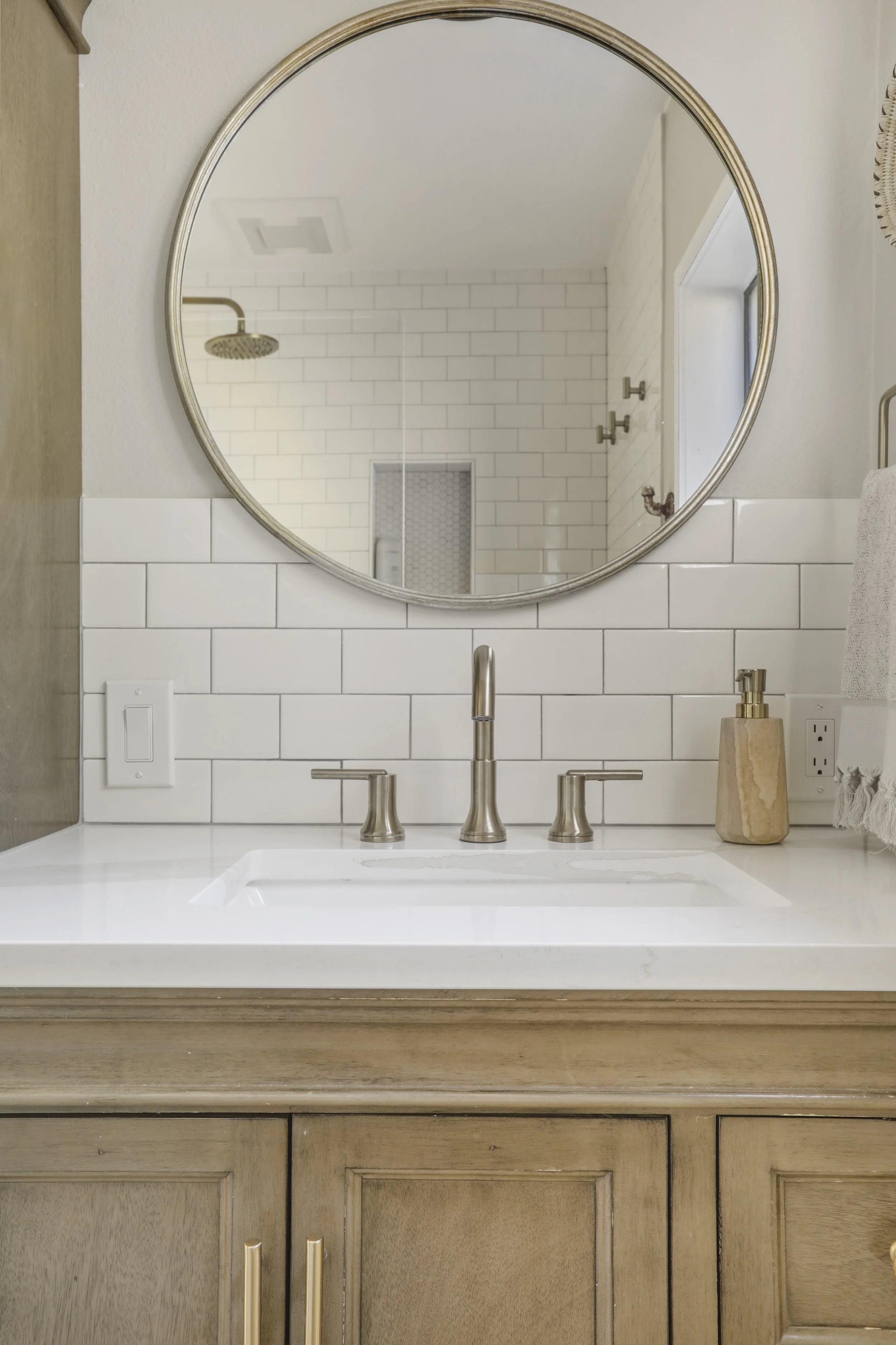 A beautifully styled bathroom vanity featuring a light wood cabinet with brass hardware, topped with a sleek white countertop and a brushed nickel three-piece faucet set. Above the vanity hangs a large round mirror with a thin metallic frame, reflect