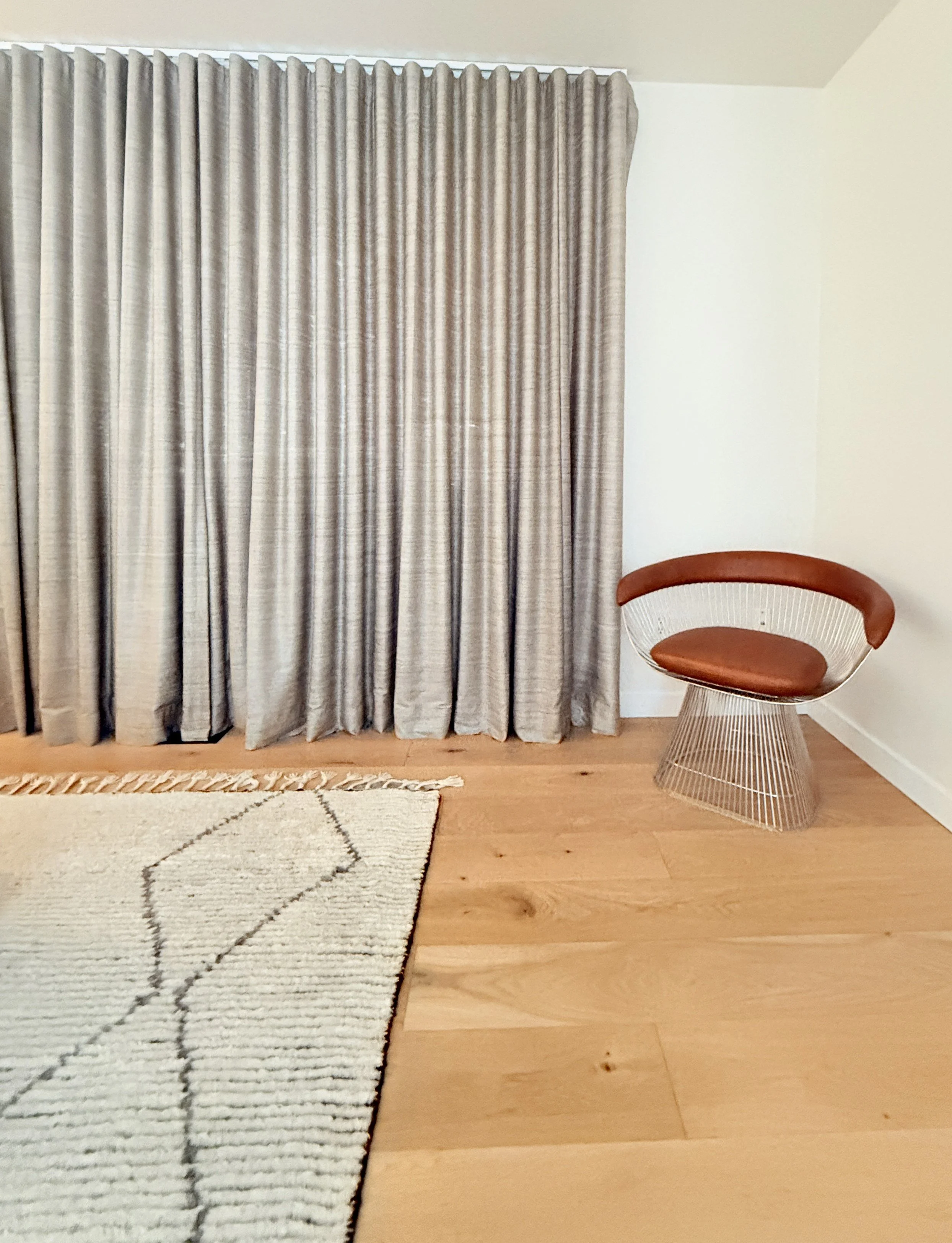 A minimalist corner of a modern room featuring light wood floors, a geometric-patterned area rug, and full-length gray curtains that add soft texture to the space. A sculptural accent chair with a wire metal base and curved wooden backrest paired wit