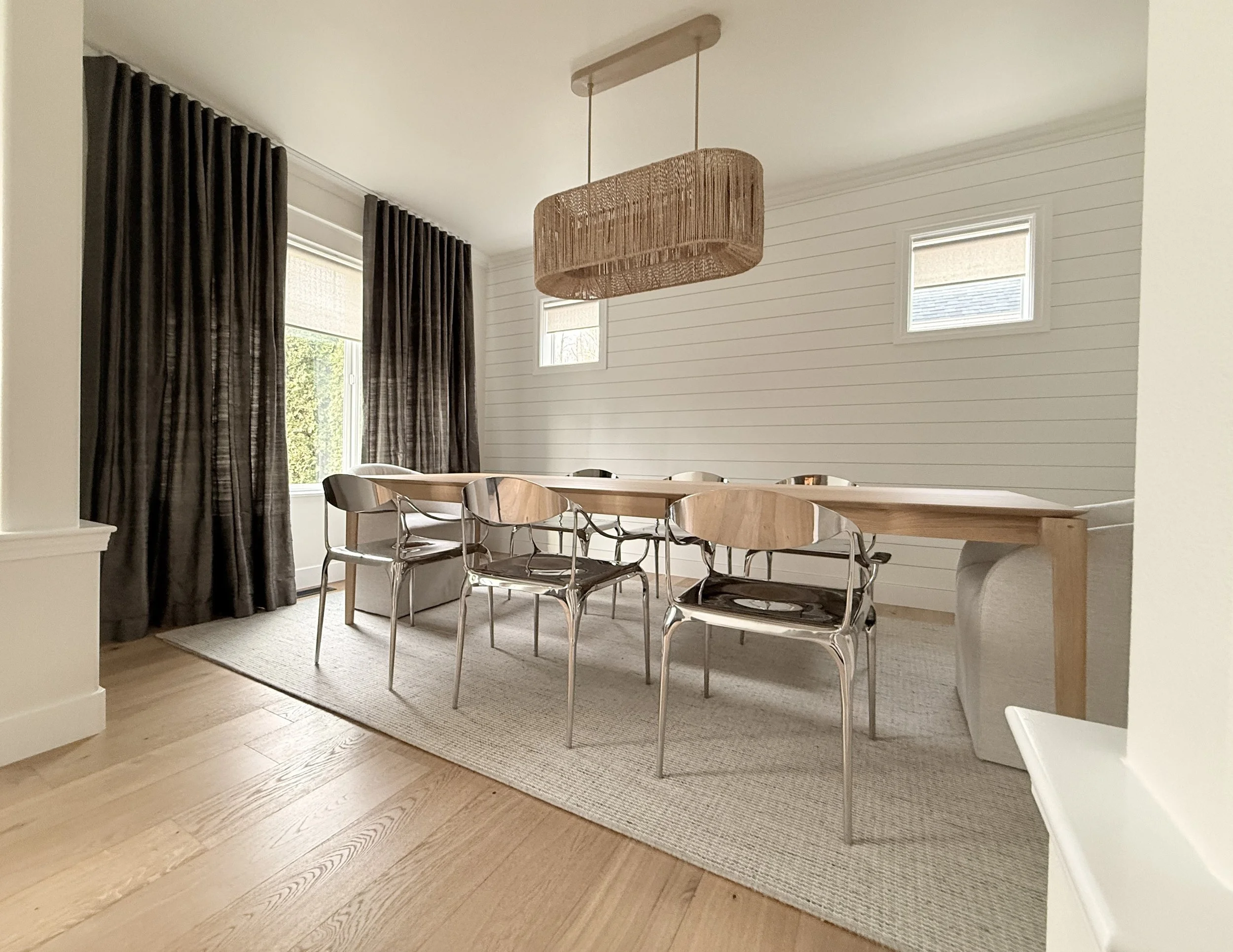 A modern dining room with a clean, airy design and natural textures. The space features a large wooden dining table surrounded by mixed-material chairs—wooden seats with chrome legs—set atop a neutral woven area rug. A woven rattan pendant light adds