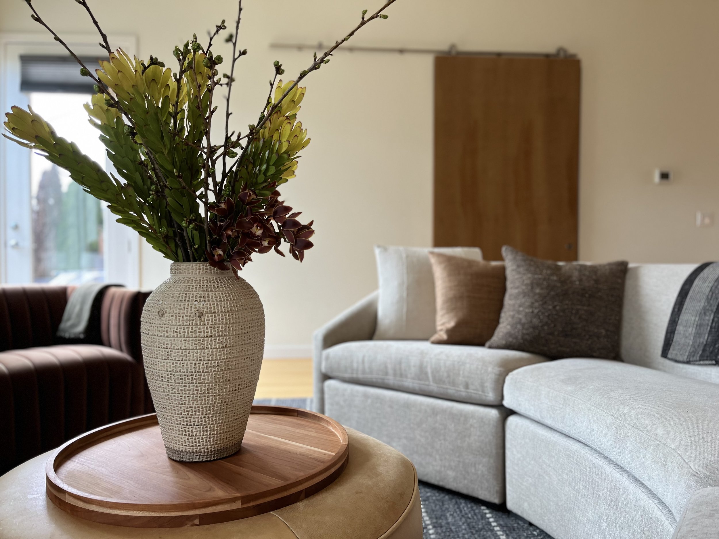 A cozy and contemporary living room vignette featuring a large round ottoman topped with a wooden tray and a textured ceramic vase filled with tall greenery and florals. In the background, a light gray sectional sofa is adorned with an assortment of 