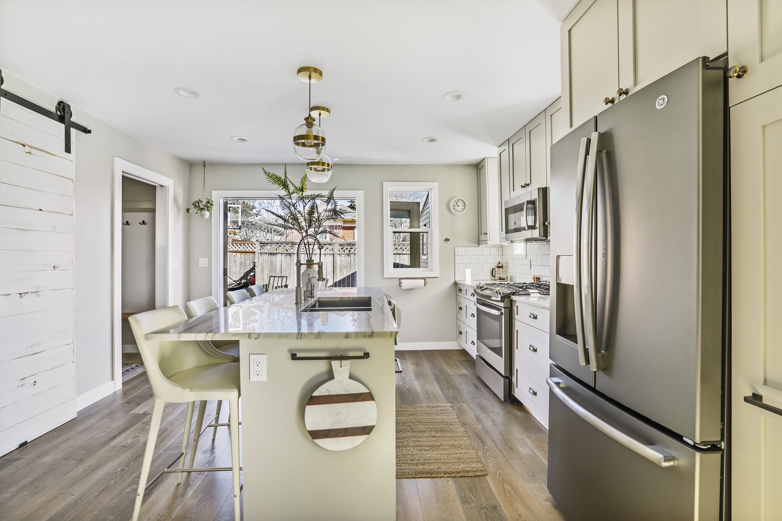 A bright and modern kitchen with a clean, open layout and stylish design elements. The space features a central island with a light gray base and a marble countertop, accompanied by sleek cream-colored bar stools. A set of three gold-accented pendant