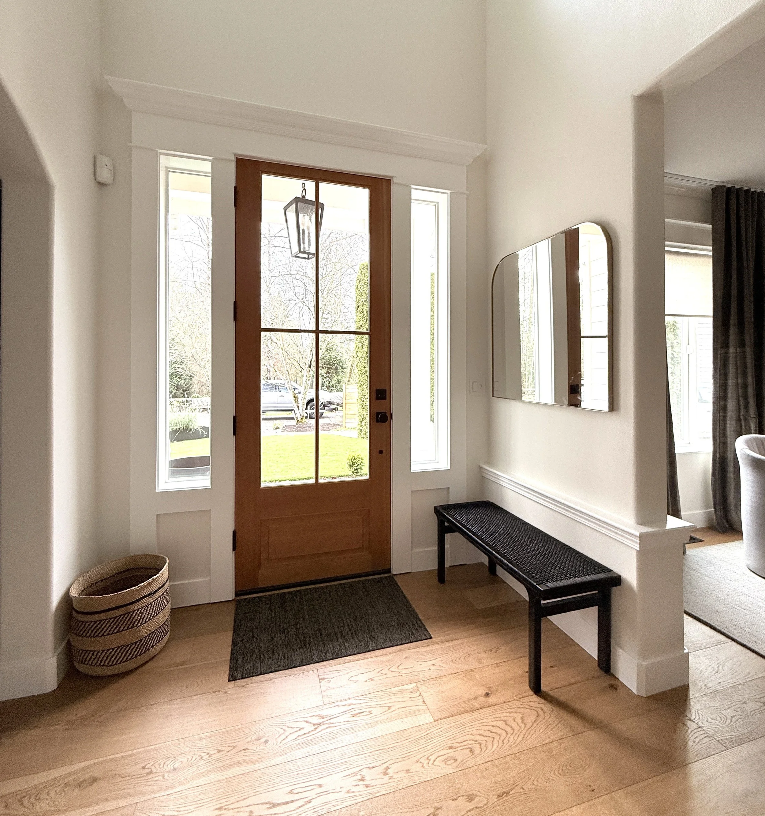 A welcoming and light-filled entryway with a natural and minimalist aesthetic. The space features a wooden front door with glass panels flanked by tall side windows, allowing abundant natural light to pour in. A black woven bench sits along the right
