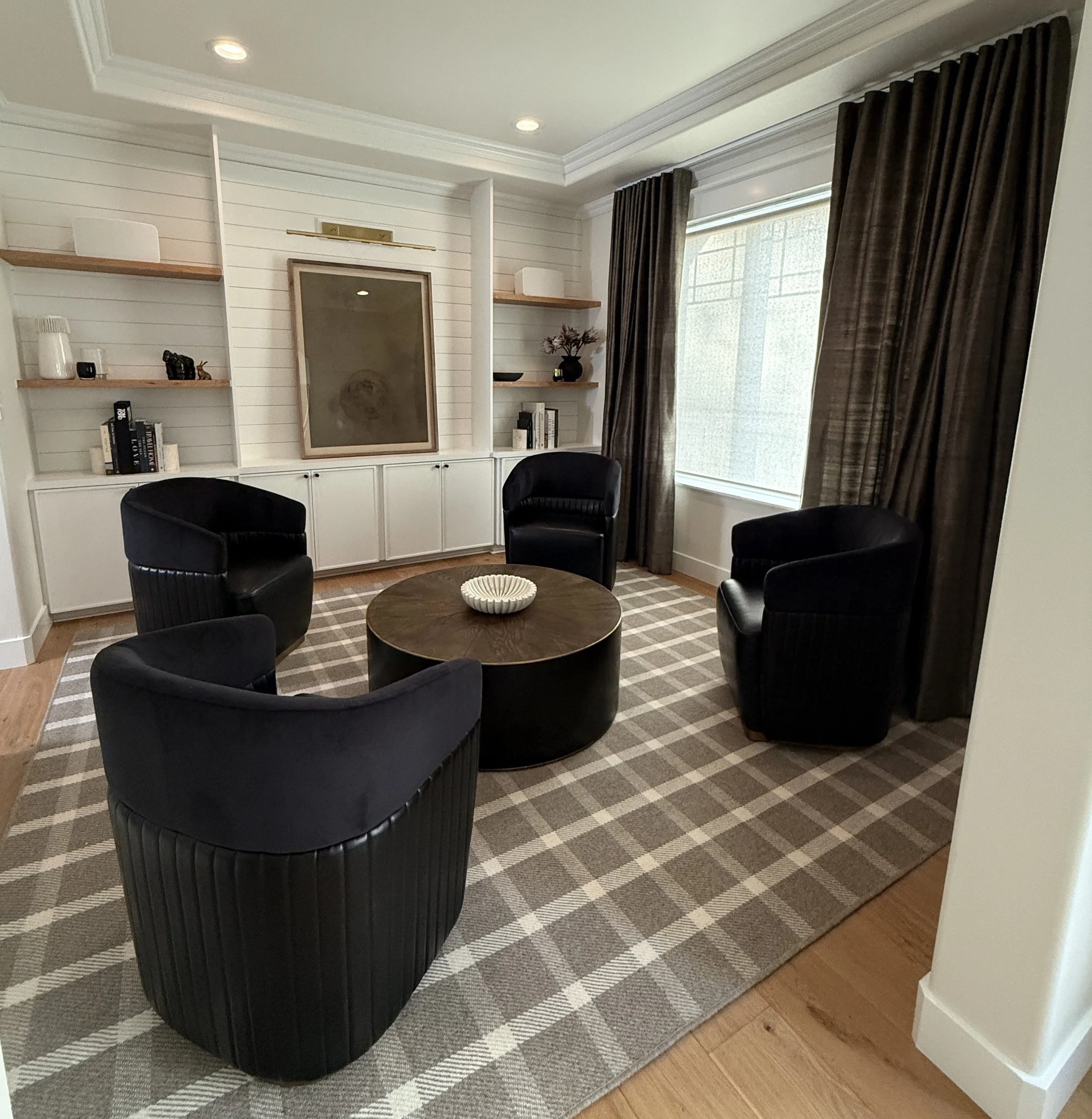 A sophisticated and intimate sitting room designed for conversation and comfort. The space features four black velvet barrel chairs arranged around a round wooden coffee table, which is topped with a sculptural white bowl. The plaid area rug in neutr