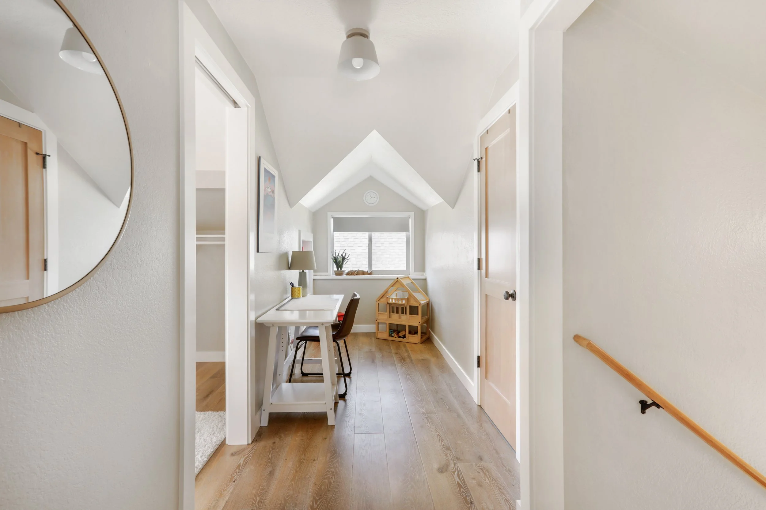 A bright and inviting attic hallway featuring a pitched ceiling and warm wood flooring. At the end of the hallway is a cozy nook with a window, a small desk with a lamp, and a wooden chair, creating a functional workspace. A dollhouse and potted plan
