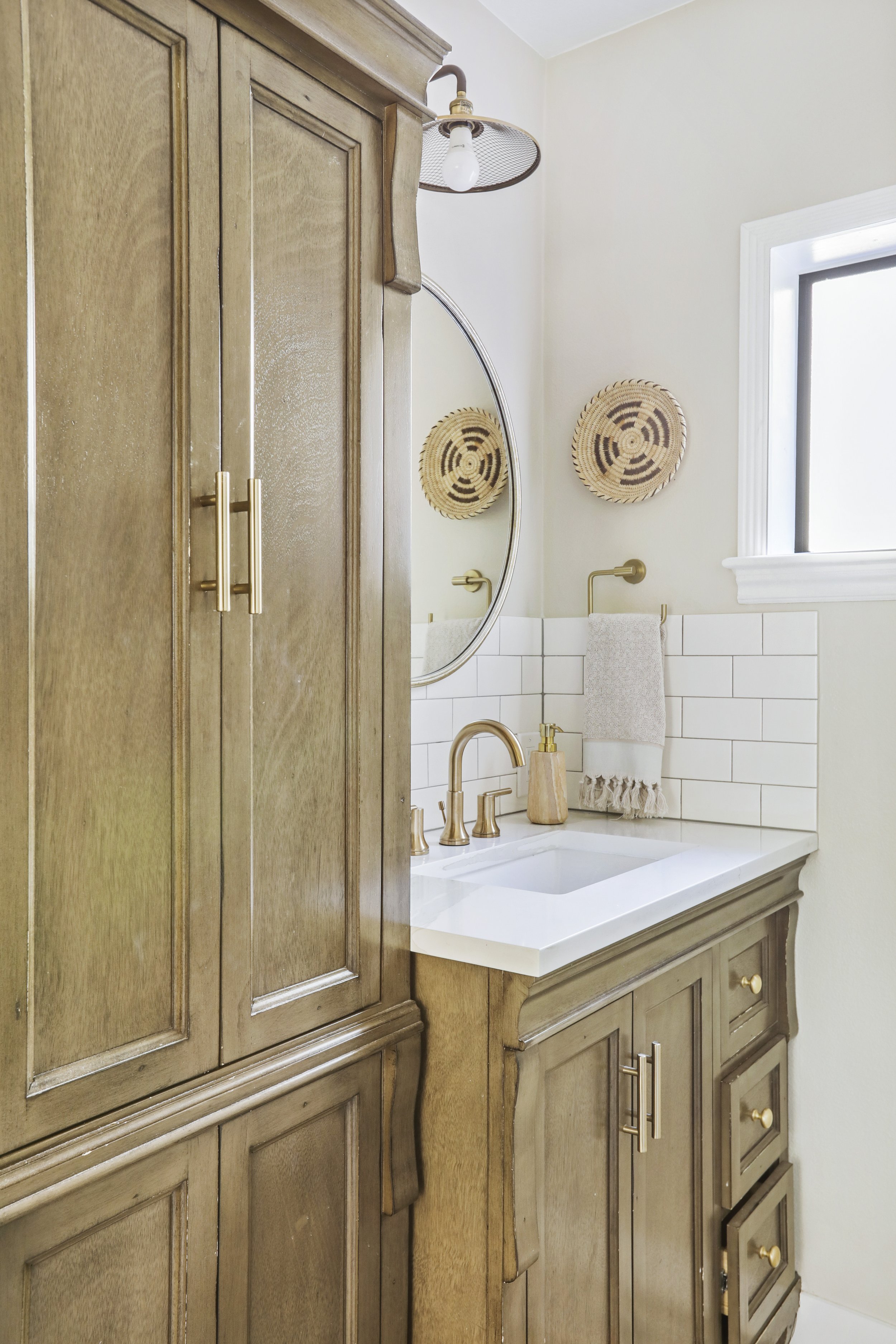 A charming and well-organized bathroom vanity area featuring warm wood cabinetry with brass hardware and a white countertop. A round mirror hangs above the sink, accompanied by a brass faucet and matching wall-mounted towel ring. The backsplash is co