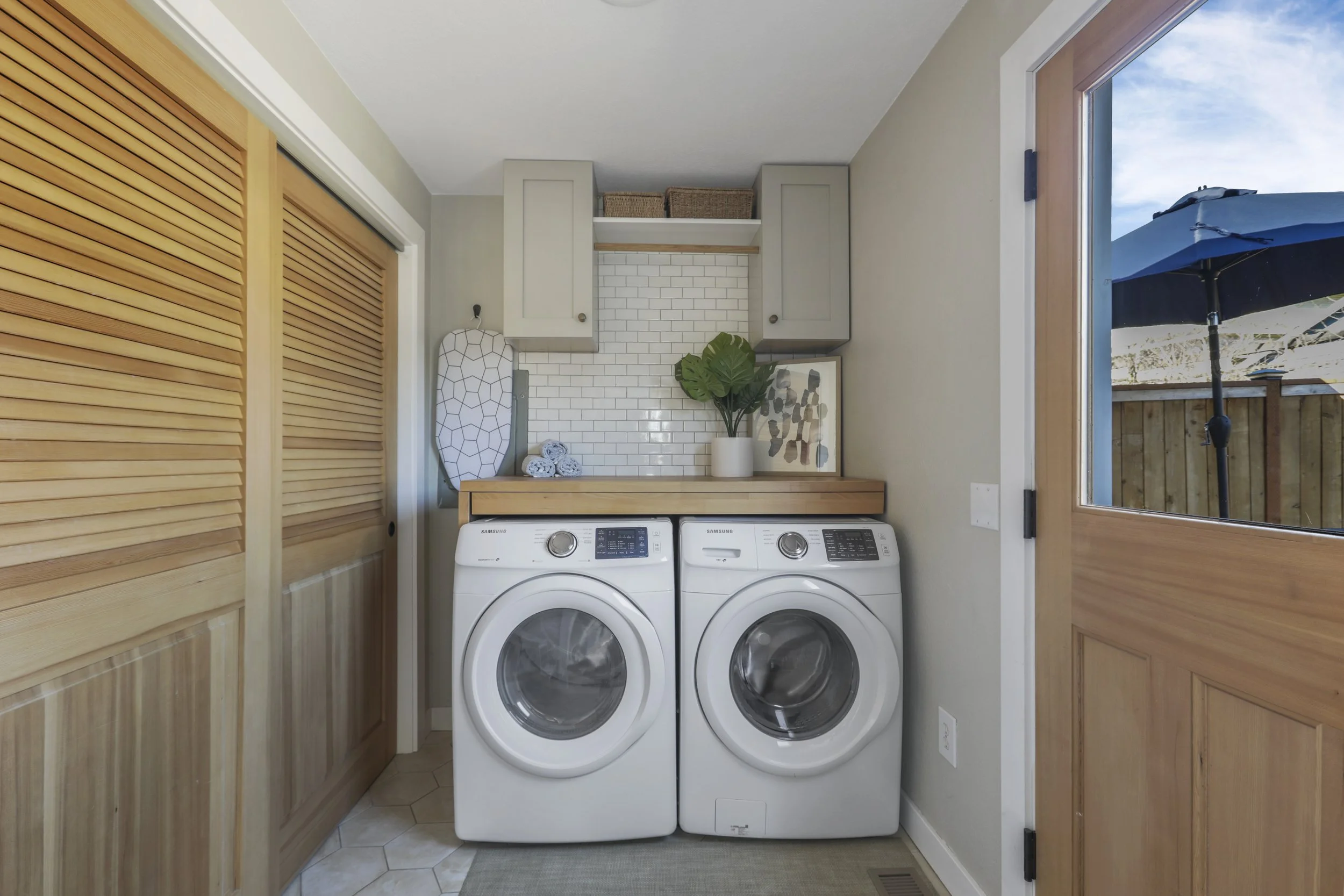 A stylish and compact laundry room featuring a front-loading washer and dryer set beneath a wood countertop that provides additional folding space. Above the machines, white subway tile with light grout serves as a clean backsplash, flanked by gray u