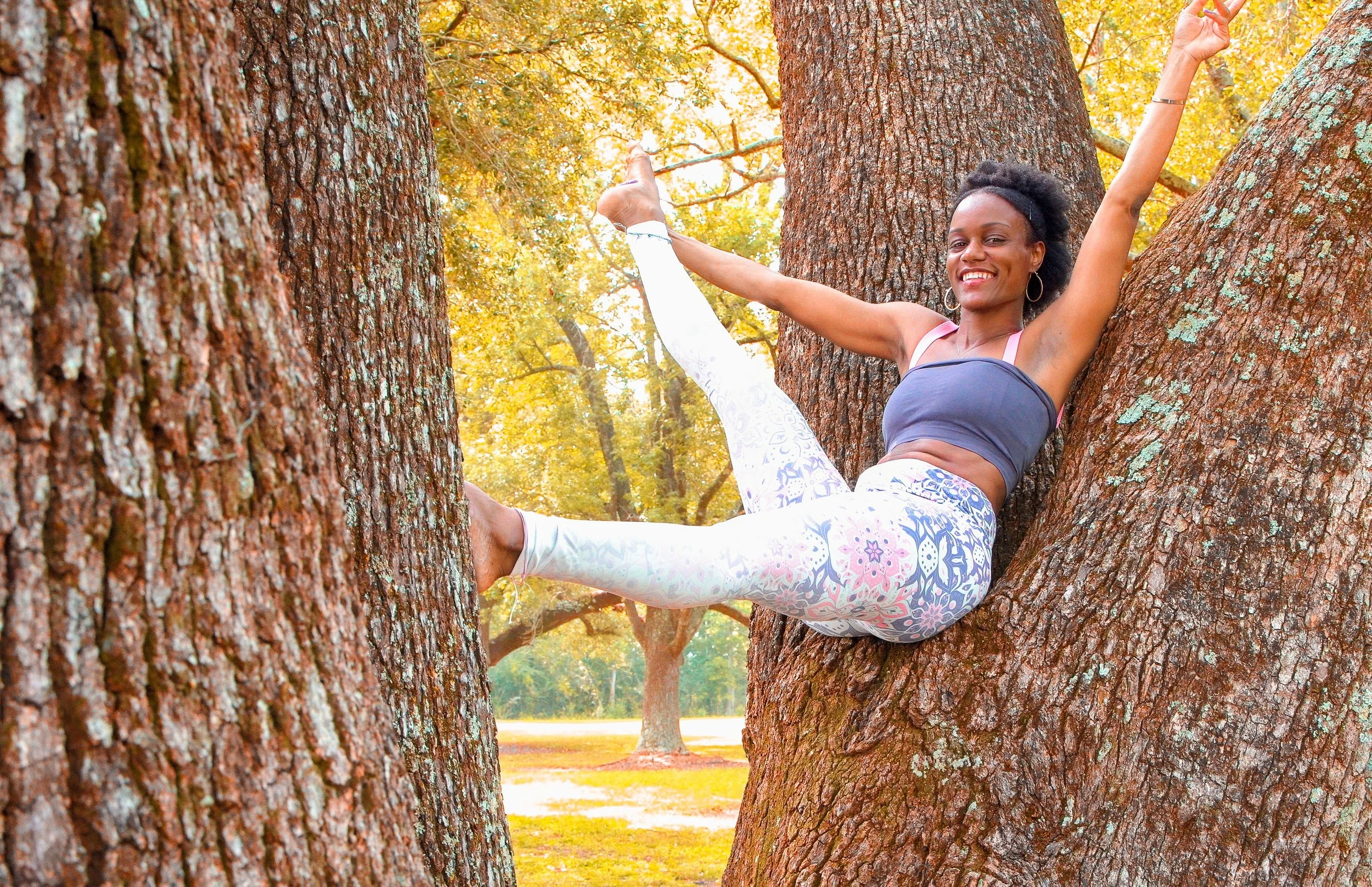 Woman smiling and sitting in a tree, extending her arms and legs in a joyful pose, surrounded by autumn trees.