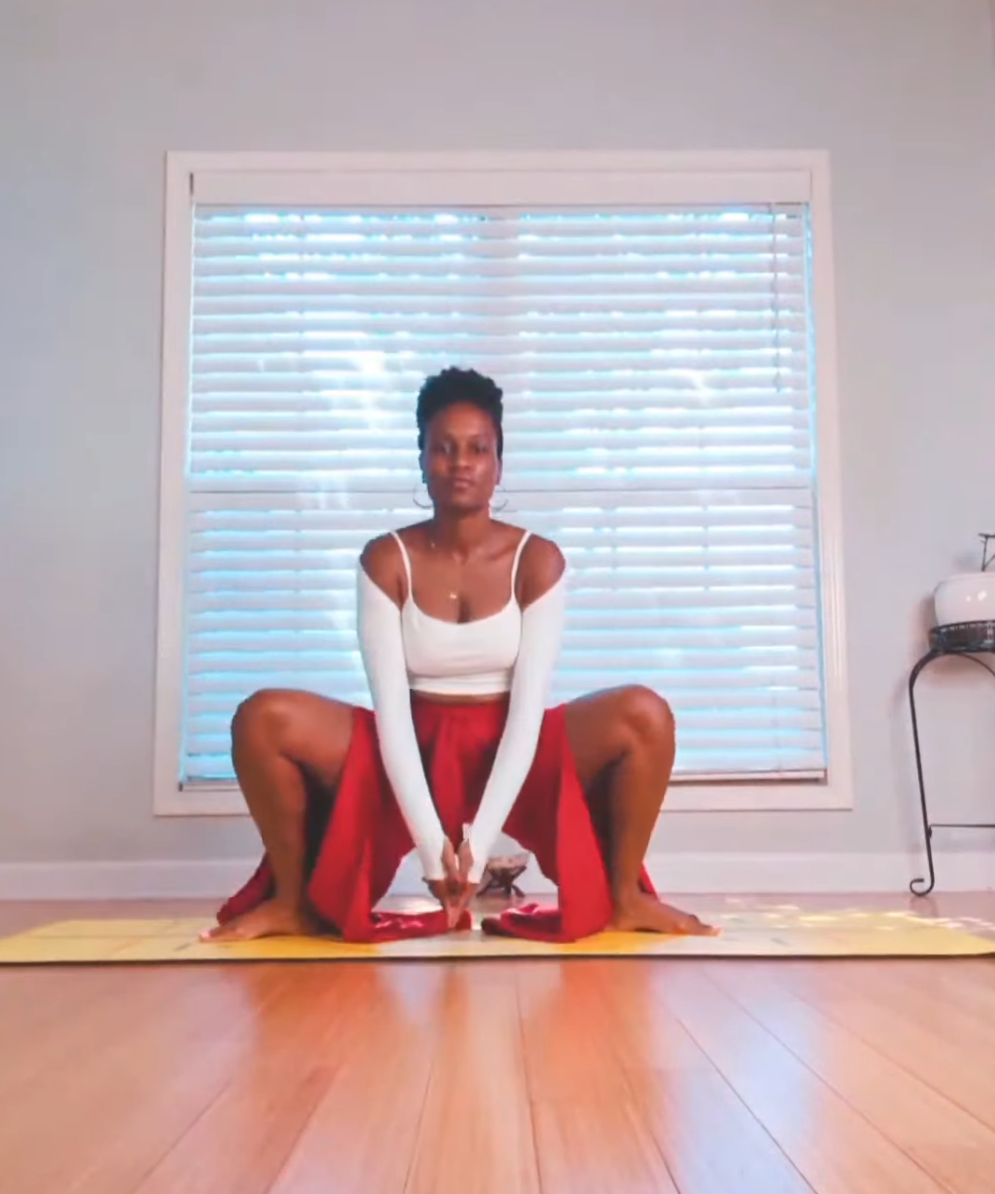 A woman practicing yoga in a room with wooden flooring, sitting on a yellow yoga mat in front of closed white blinds on a window, dressed in a white top and red pants.