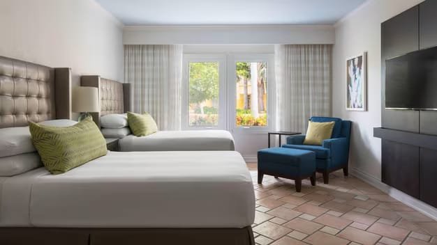 Hotel room with two beds, a blue armchair, and a large window with curtains.
