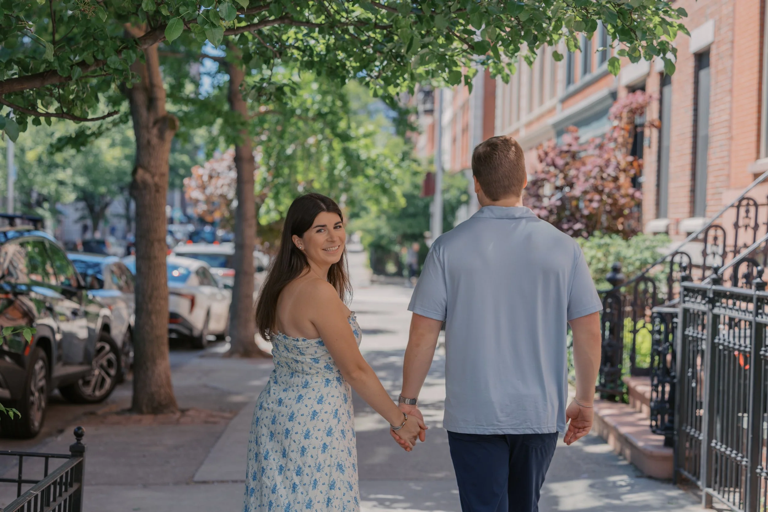 A couple holding hands walking on a residential sidewalk, with the woman smiling at the camera and the man facing away, surrounded by trees and brick row houses.
