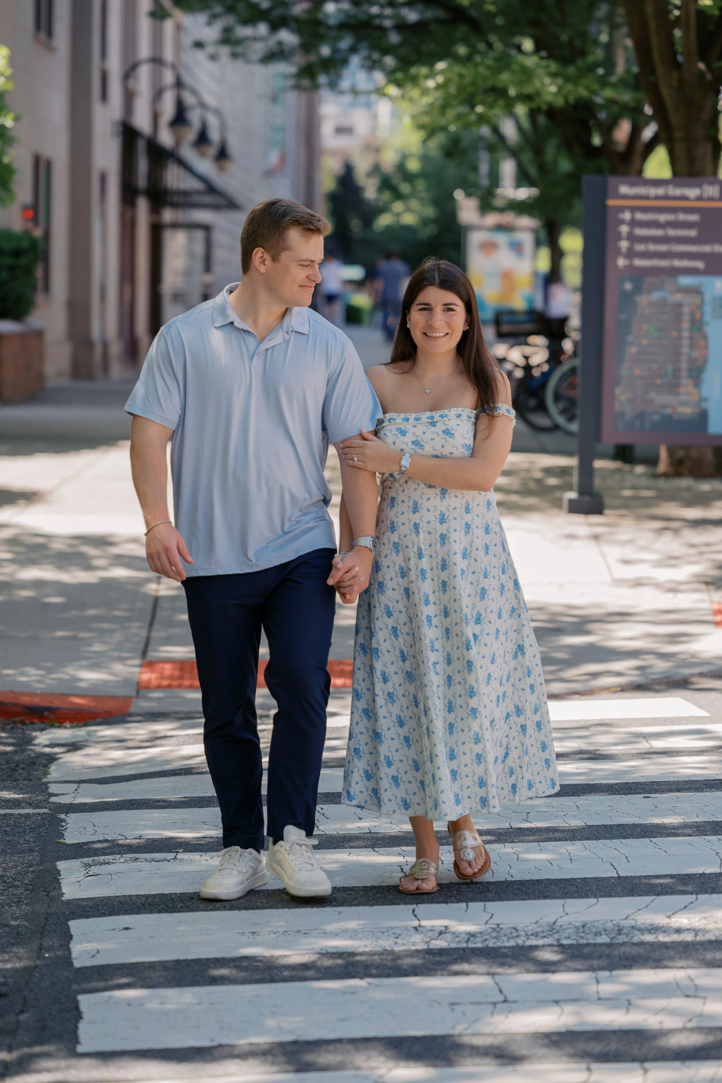 A young man and woman holding hands walking across a crosswalk in an urban area on a sunny day. The woman is smiling and wearing a floral dress and sandals, while the man is smiling and wearing a light blue shirt, dark pants, and white sneakers.