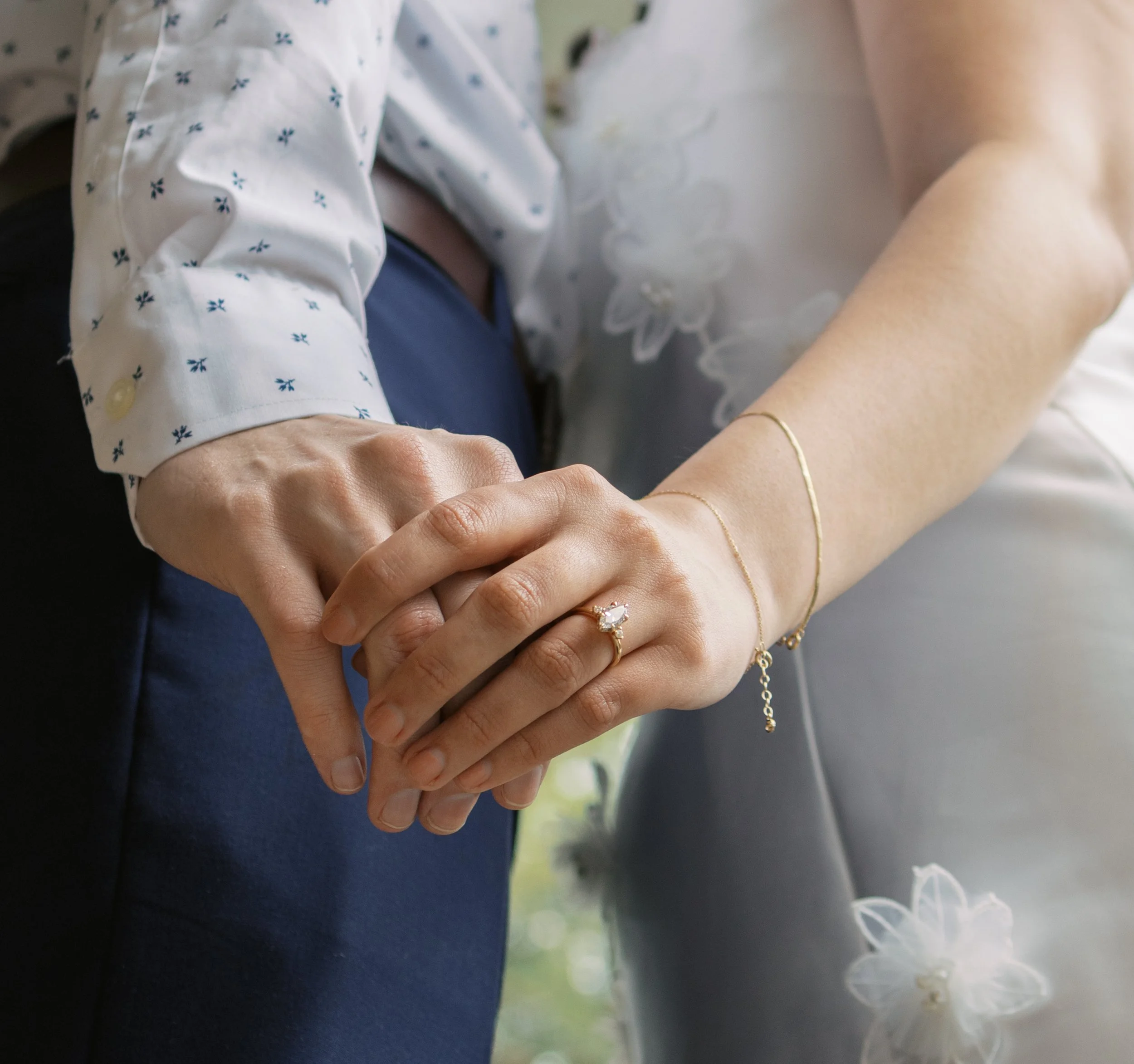 Close-up of two people holding hands, one wearing a wedding ring and the other a gold bracelet, with a blurred white dress in the background.