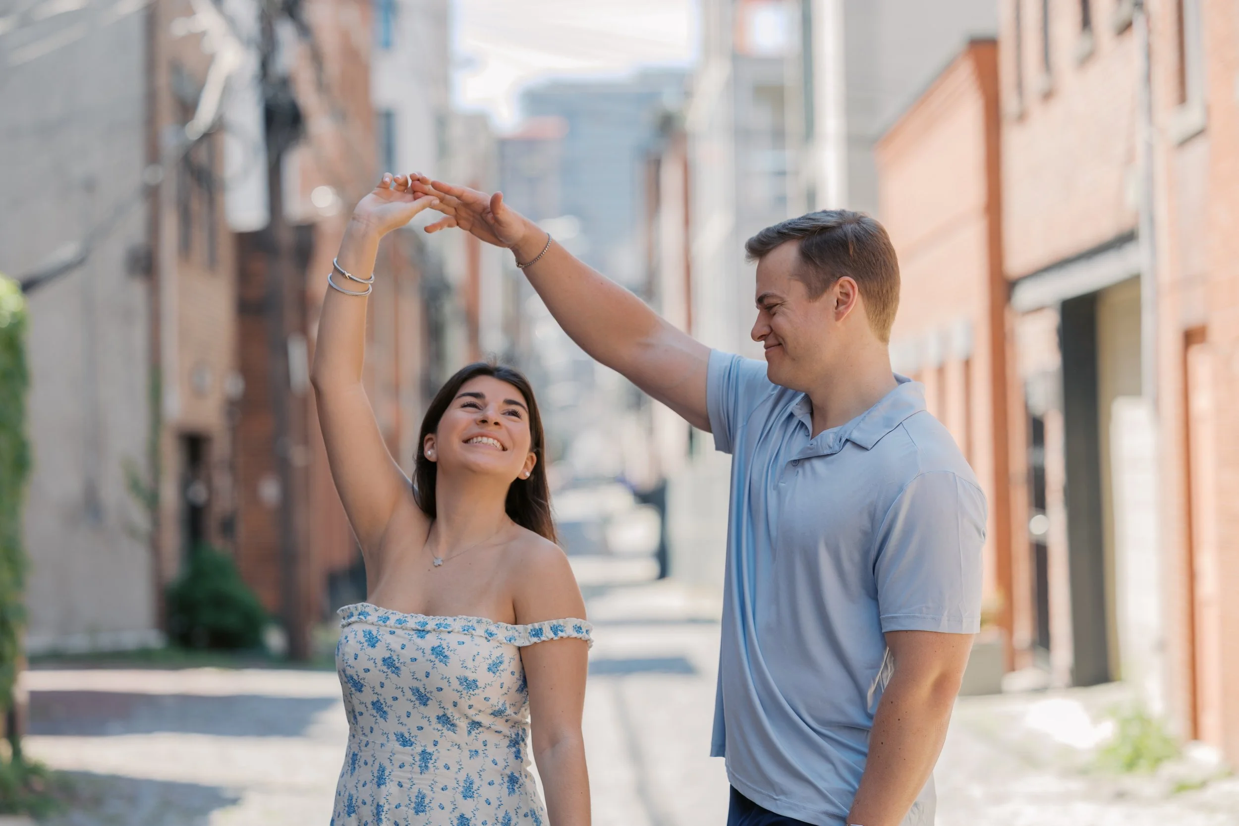 A young woman and a young man dancing and smiling on a city street, with the man raising his hand to hold the woman's hand.