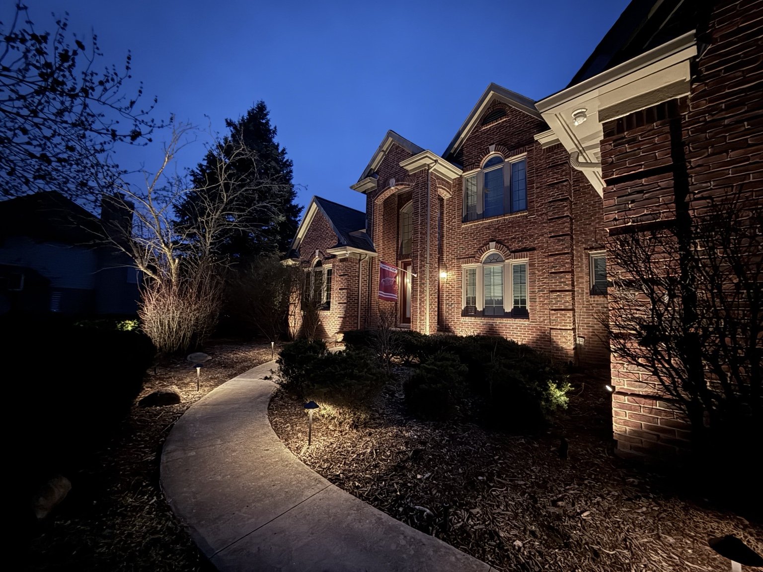 A brick house at dusk with outdoor lighting illuminating the front yard and walkway, trees, and shrubs.