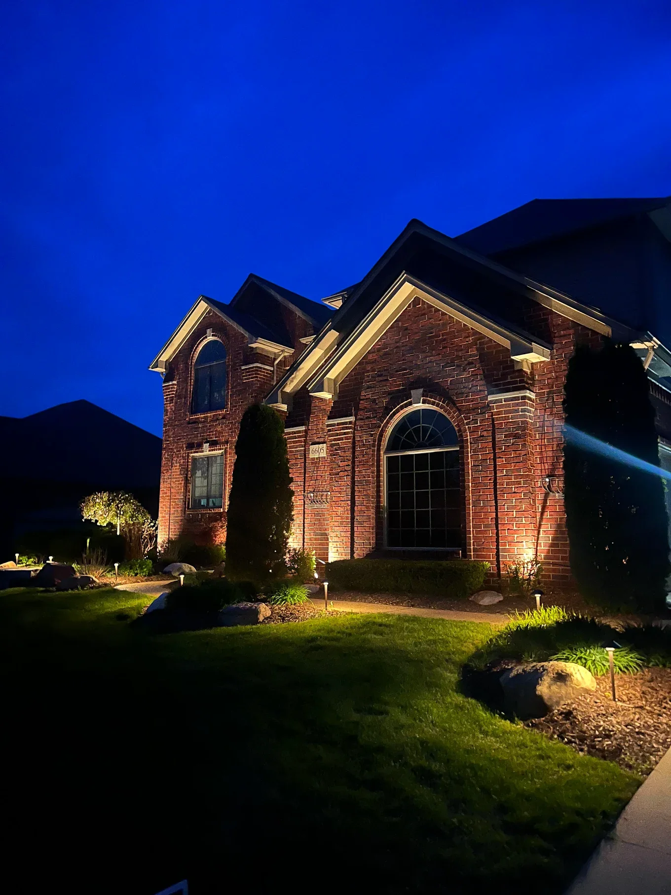 A brick house illuminated at night with landscaping lights, two tall bushes, and a well-maintained lawn.