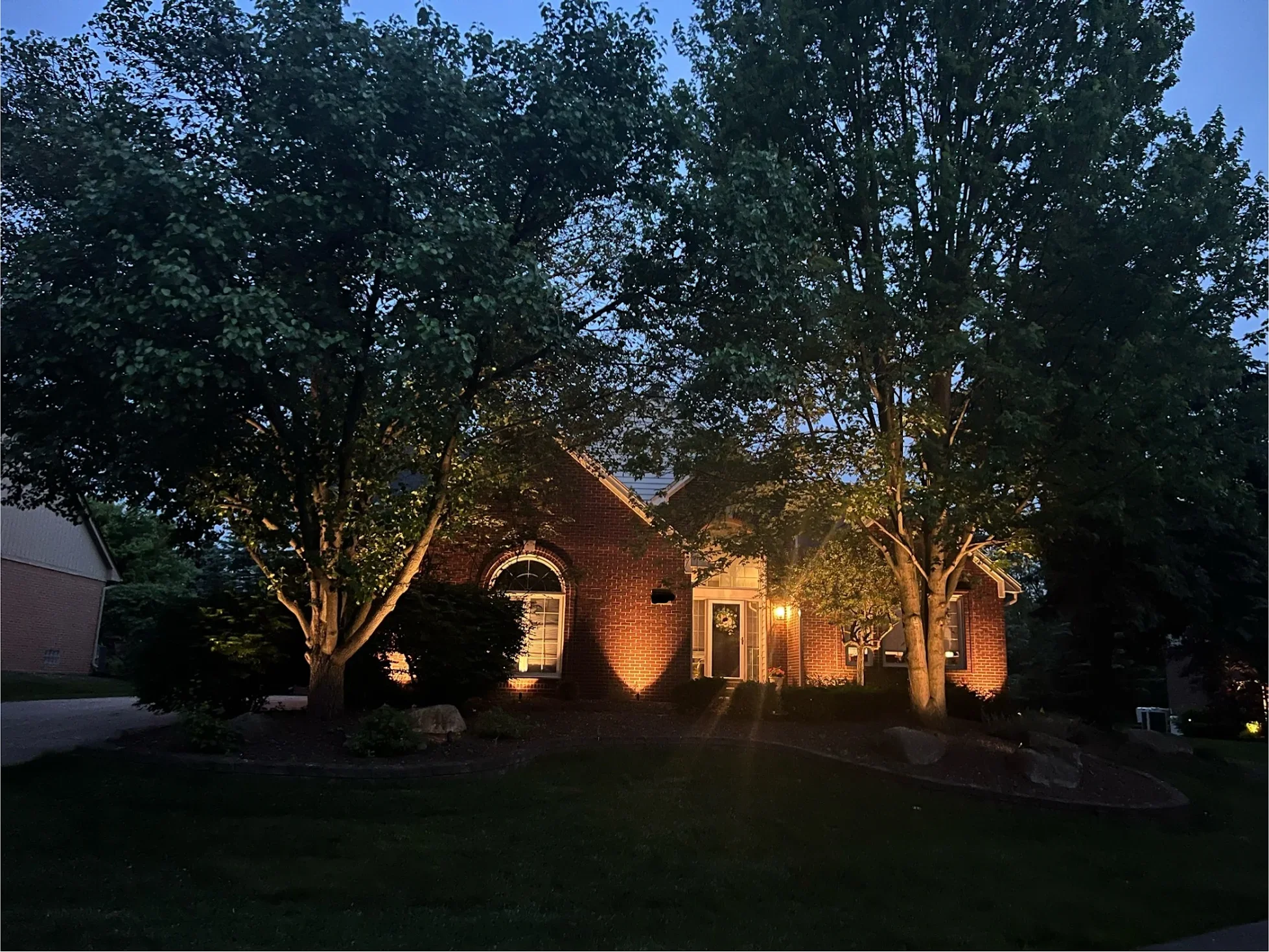 A brick house with a front porch and large arched windows, illuminated with outdoor lighting, surrounded by trees and greenery at dusk.