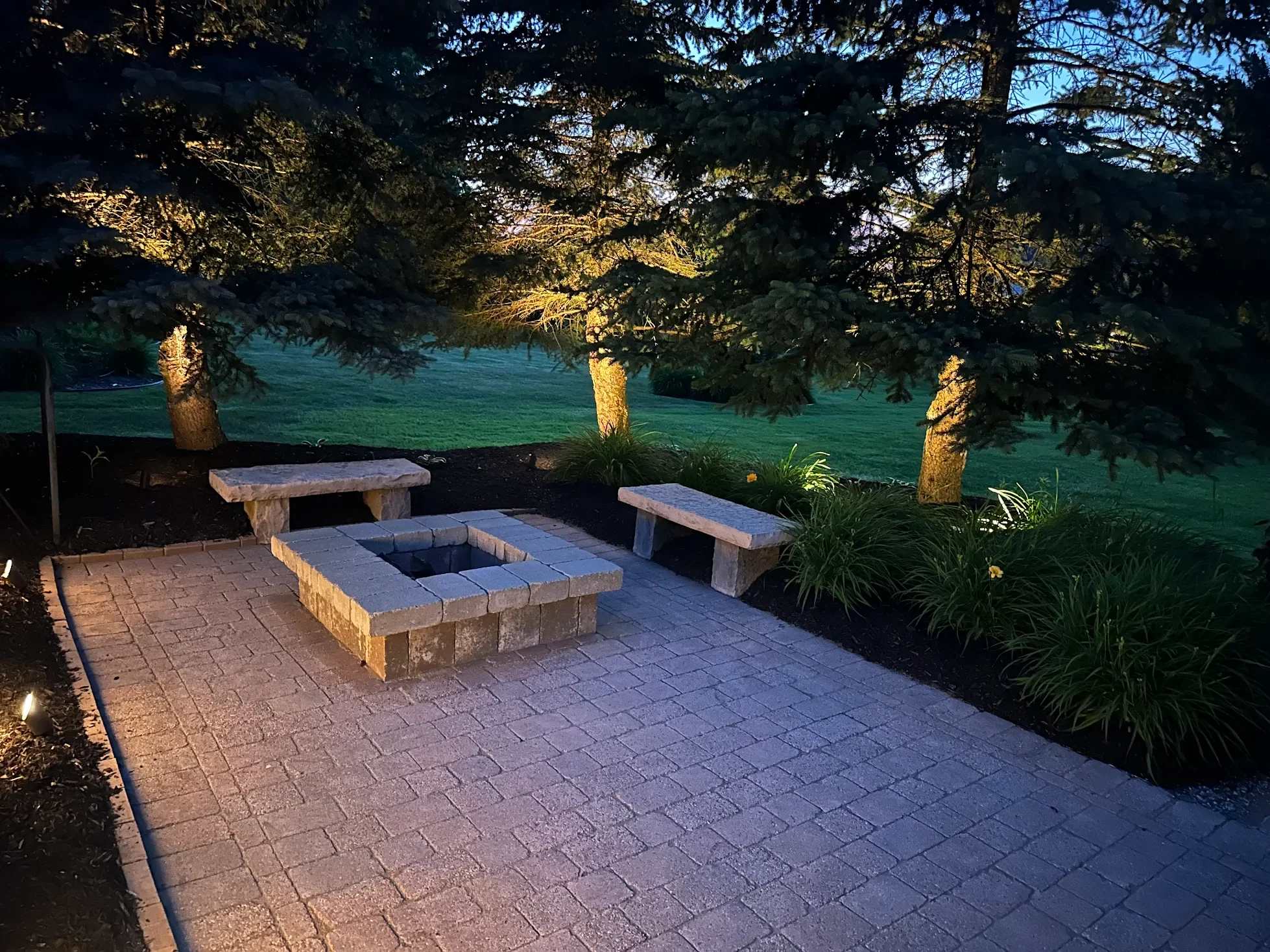 Nighttime outdoor scene showing a brick-paved patio with a stone fire pit, two stone benches, and landscaped bushes with yellow flowers, shaded by large trees.