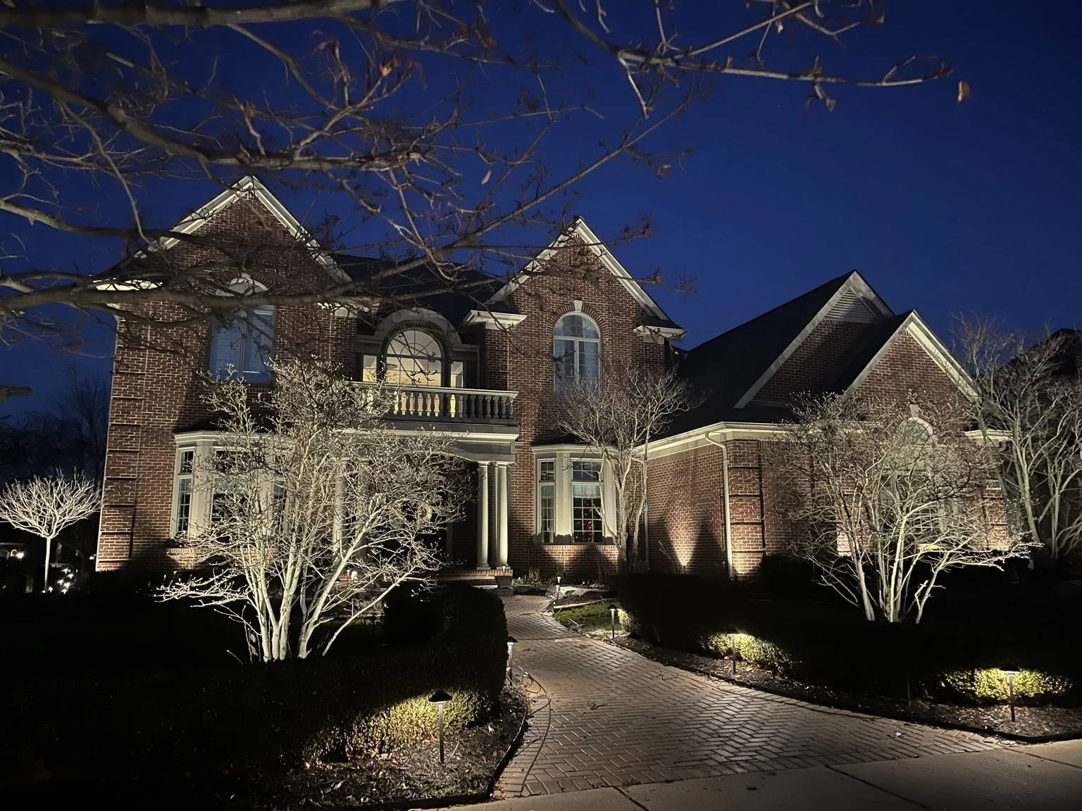 A large brick house illuminated at night with trees and bushes in the front yard, a brick pathway, and a dark blue sky.