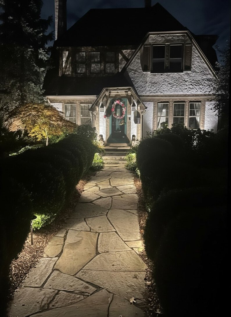 A house at night with a stone walkway leading to the front door, decorated with a Christmas wreath, with outdoor lighting illuminating the house and bushes.