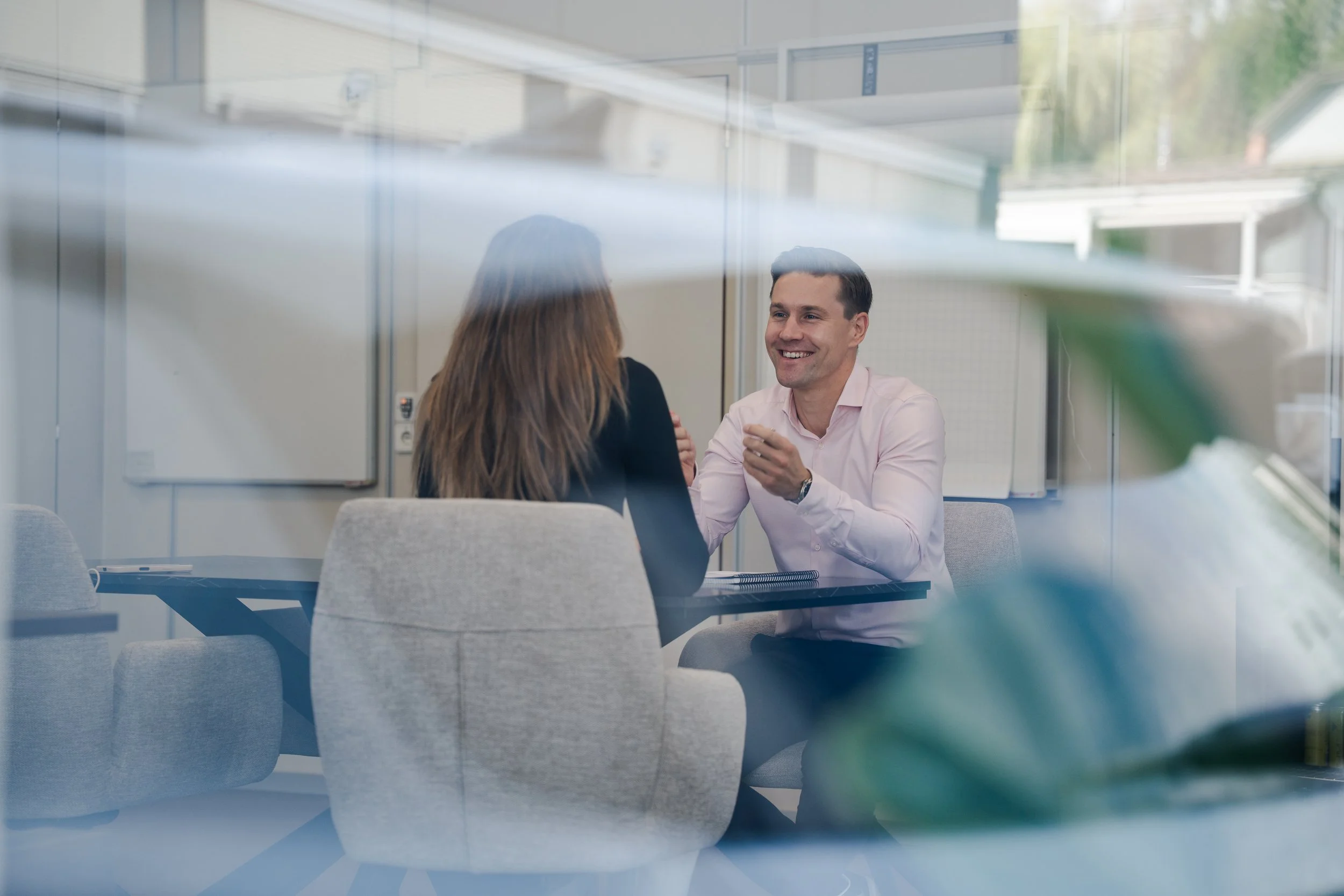 Zwei Personen im Gespräch, eine lächelnde Frau und ein Mann, sitzen an einem Konferenztisch in einem modernen Büro, durch eine Glasscheibe betrachtet.